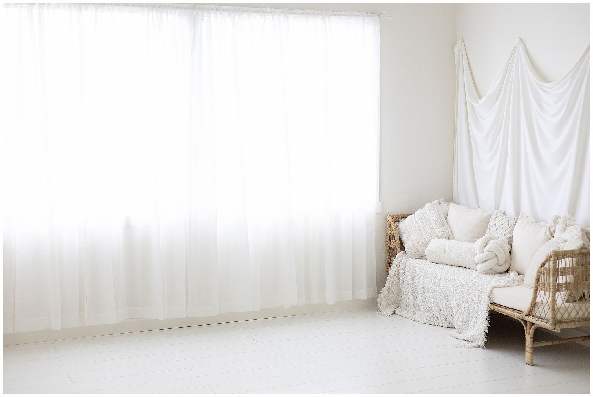Bright white photography studio with rattan sofa, white drape backdrop, and sheer curtains filtering natural light.