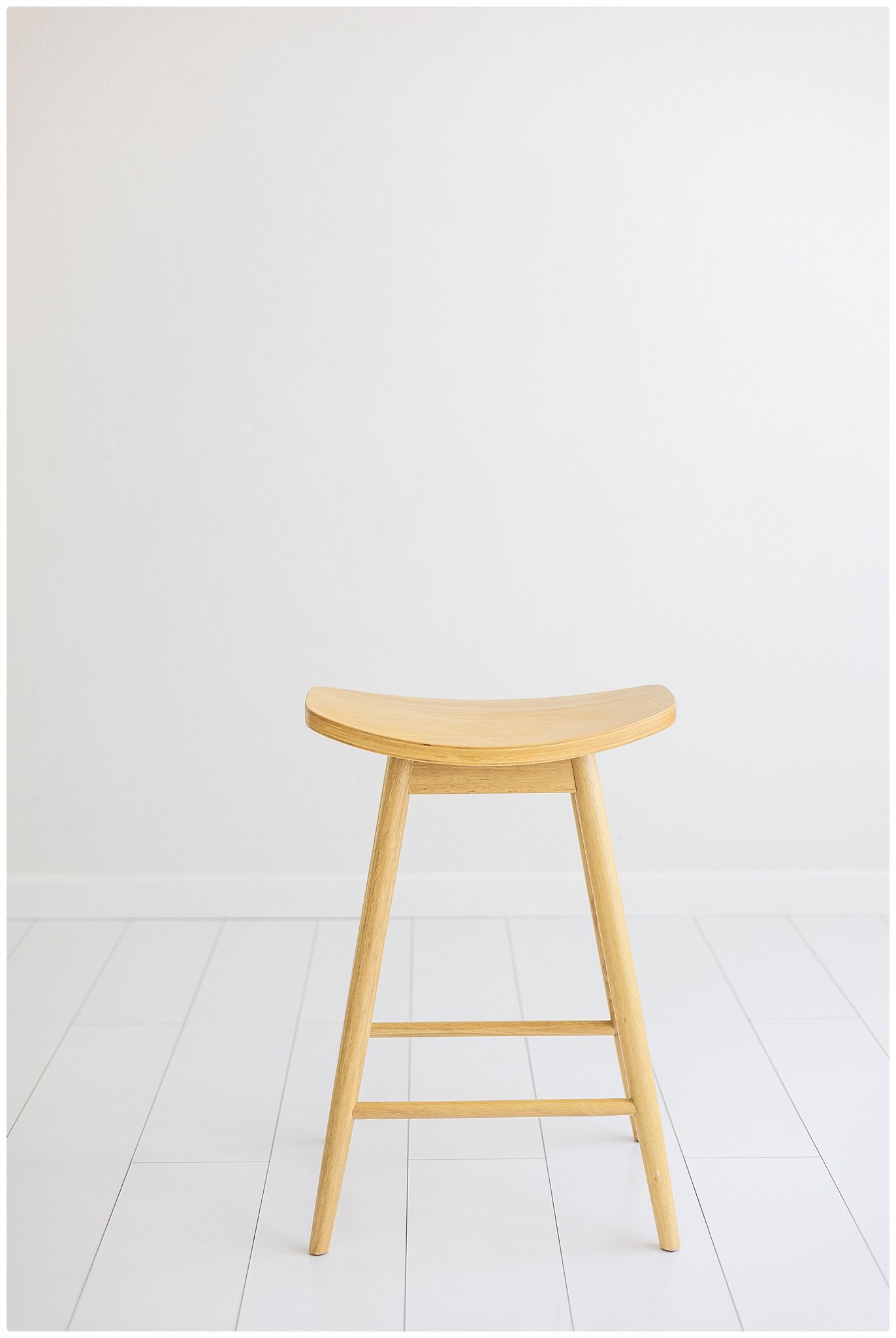 Natural oak wooden stool with curved saddle seat and four legs on white floor.