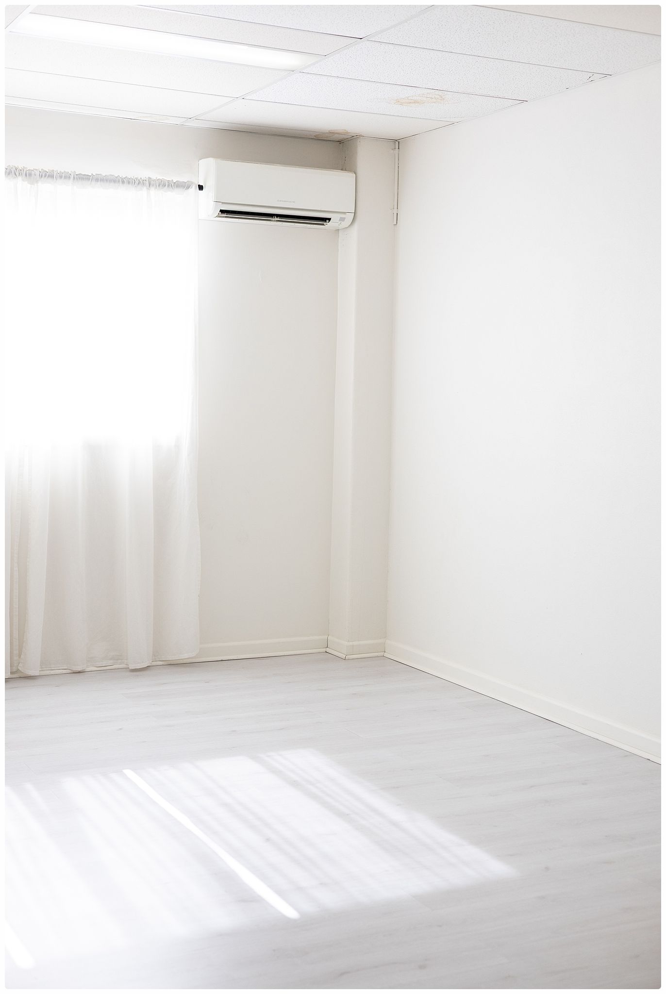 Bright minimalist white room with hardwood floors, sheer curtains, and a wall-mounted air conditioning unit.