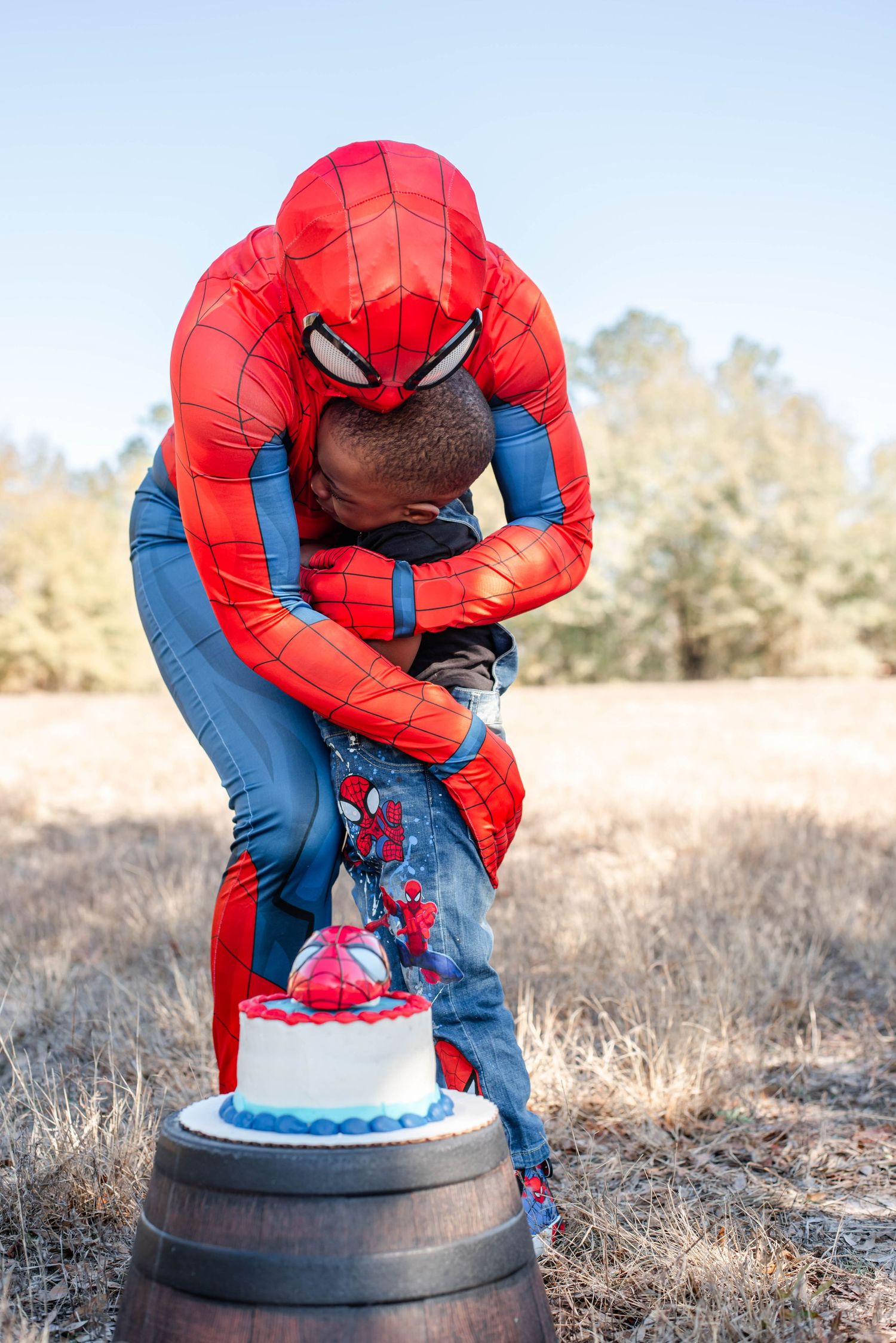 Spider-Man costume figure leans over a birthday cake decorated in red and blue on an outdoor wooden surface.