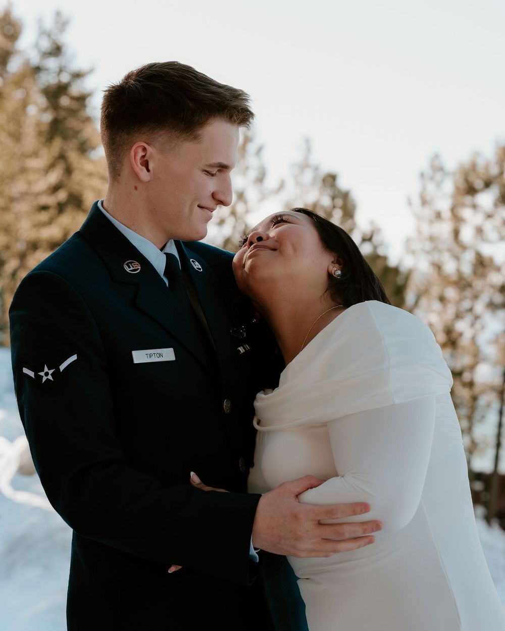 A winter wedding couple shares intimate moments in the snow with evergreen trees in the background.