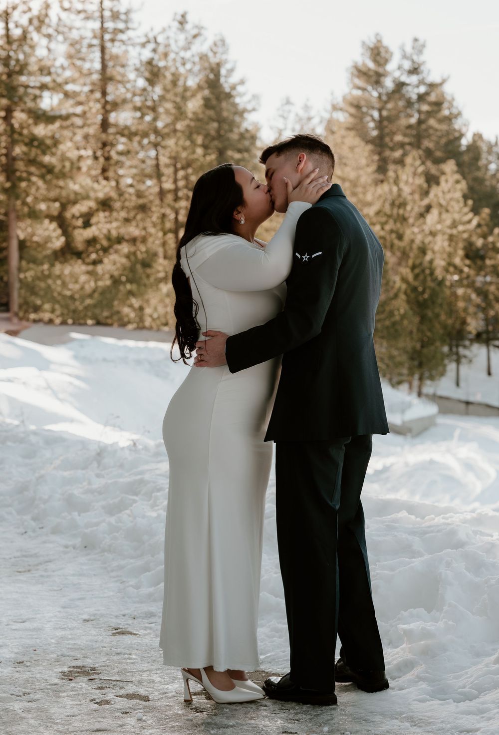 A romantic winter wedding couple shares intimate moments in a snowy forest setting with tall pine trees in the background.