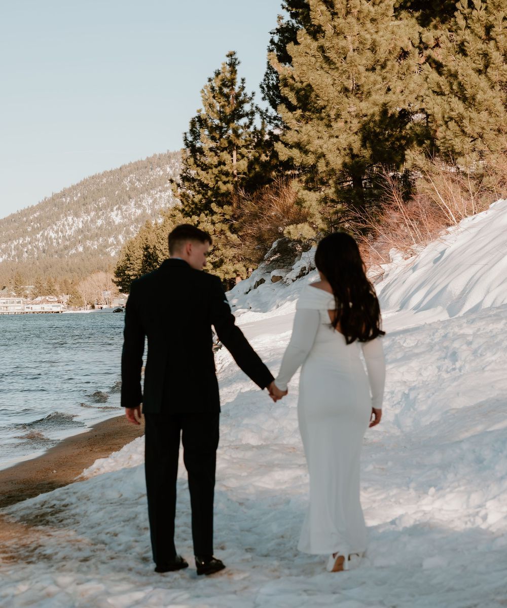 A couple walks hand in hand along a snowy winter shoreline with mountains and evergreen trees in the background.