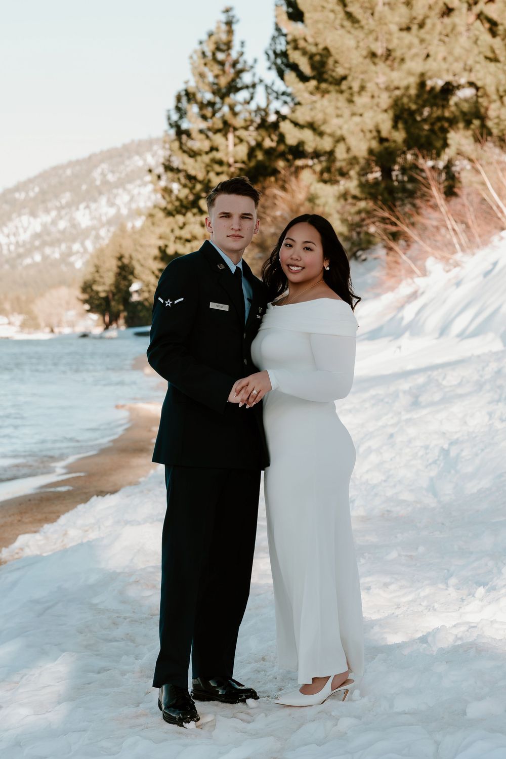 A couple in formal attire poses together on a snowy beach with mountains and pine trees in the background.