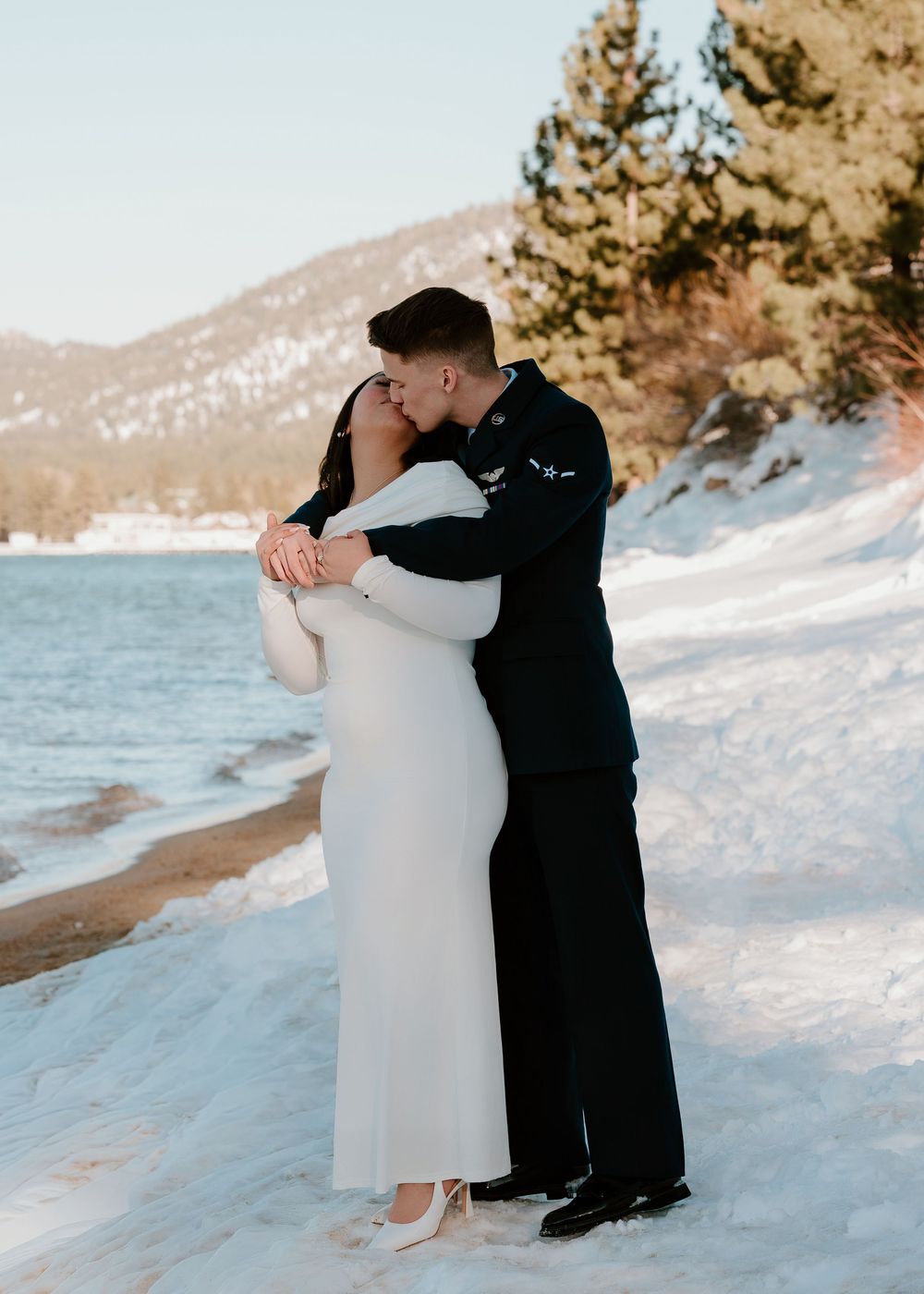 A couple embraces on a snowy beach with mountains and water in the background during winter.