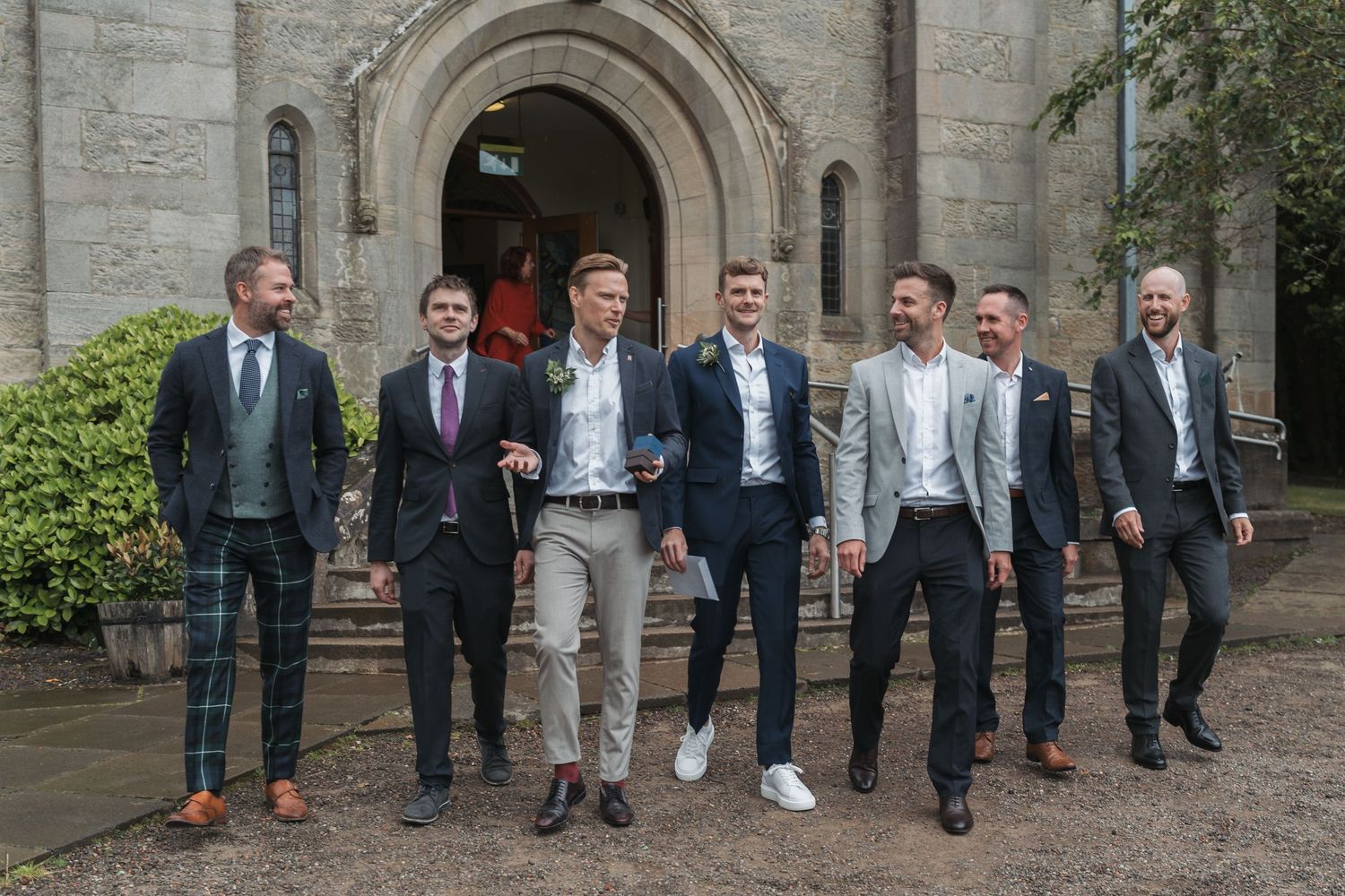 A group of groomsmen in navy suits and brown shoes pose for wedding photos in front of a stone building.