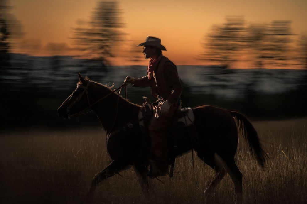 Silhouetted cowboy riding horseback against a dramatic sunset with motion-blurred trees in the background.