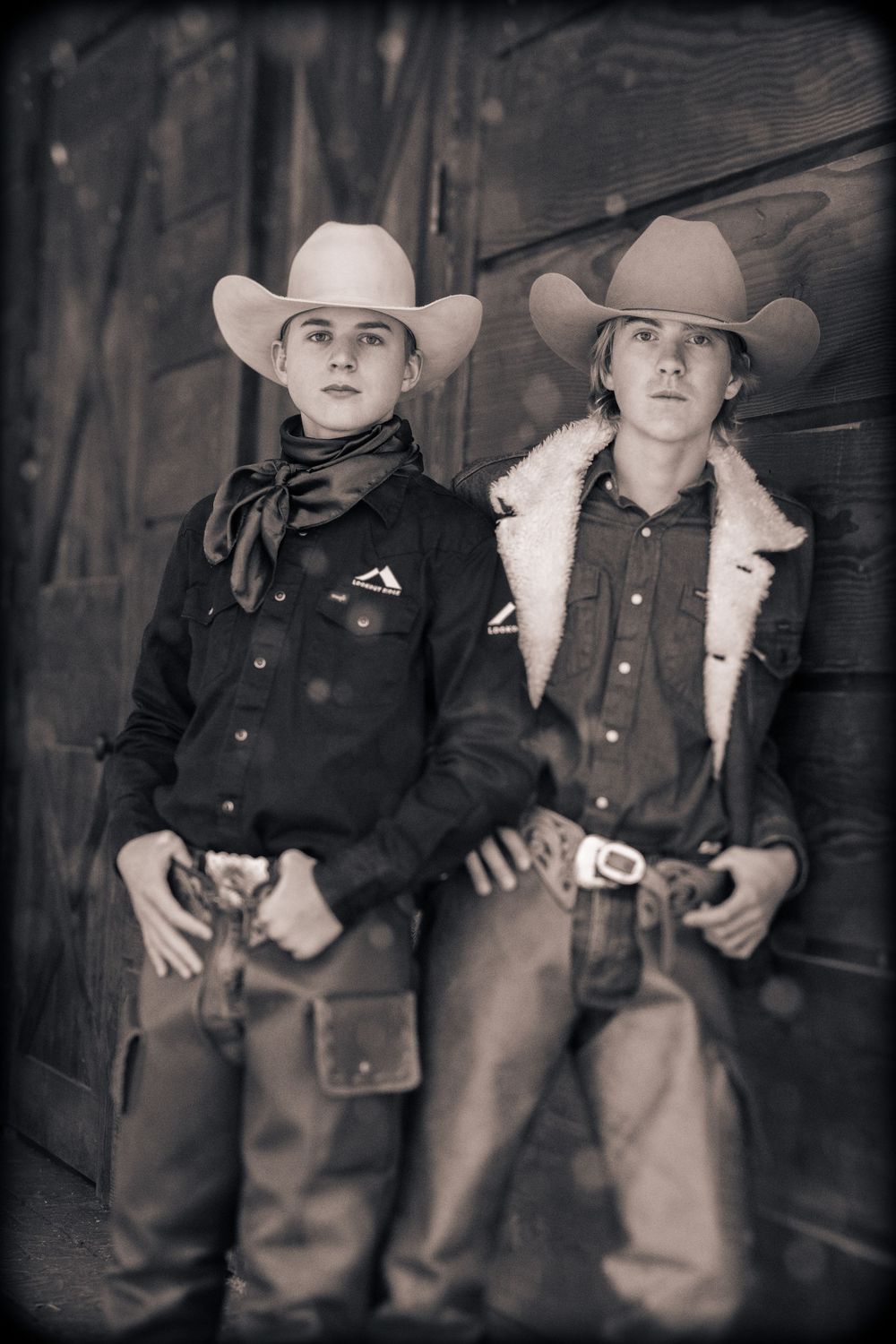 Two young cowboys in cowboy hats and western attire pose against a rustic wooden wall in a vintage-style photograph.
