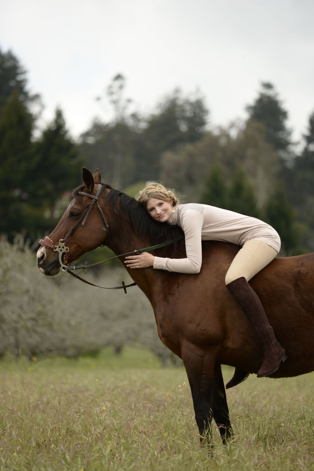 A person in white clothing lies gracefully across a brown horse's back in a grassy field with trees in the background.