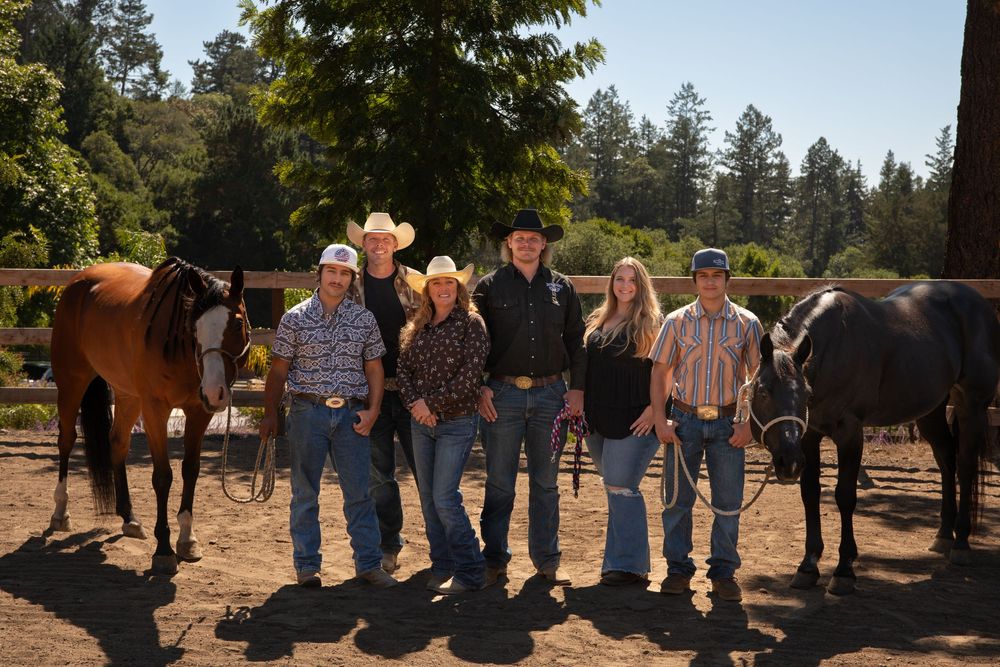 A group of people gather around horses on a sunny day at a ranch or outdoor equestrian facility.