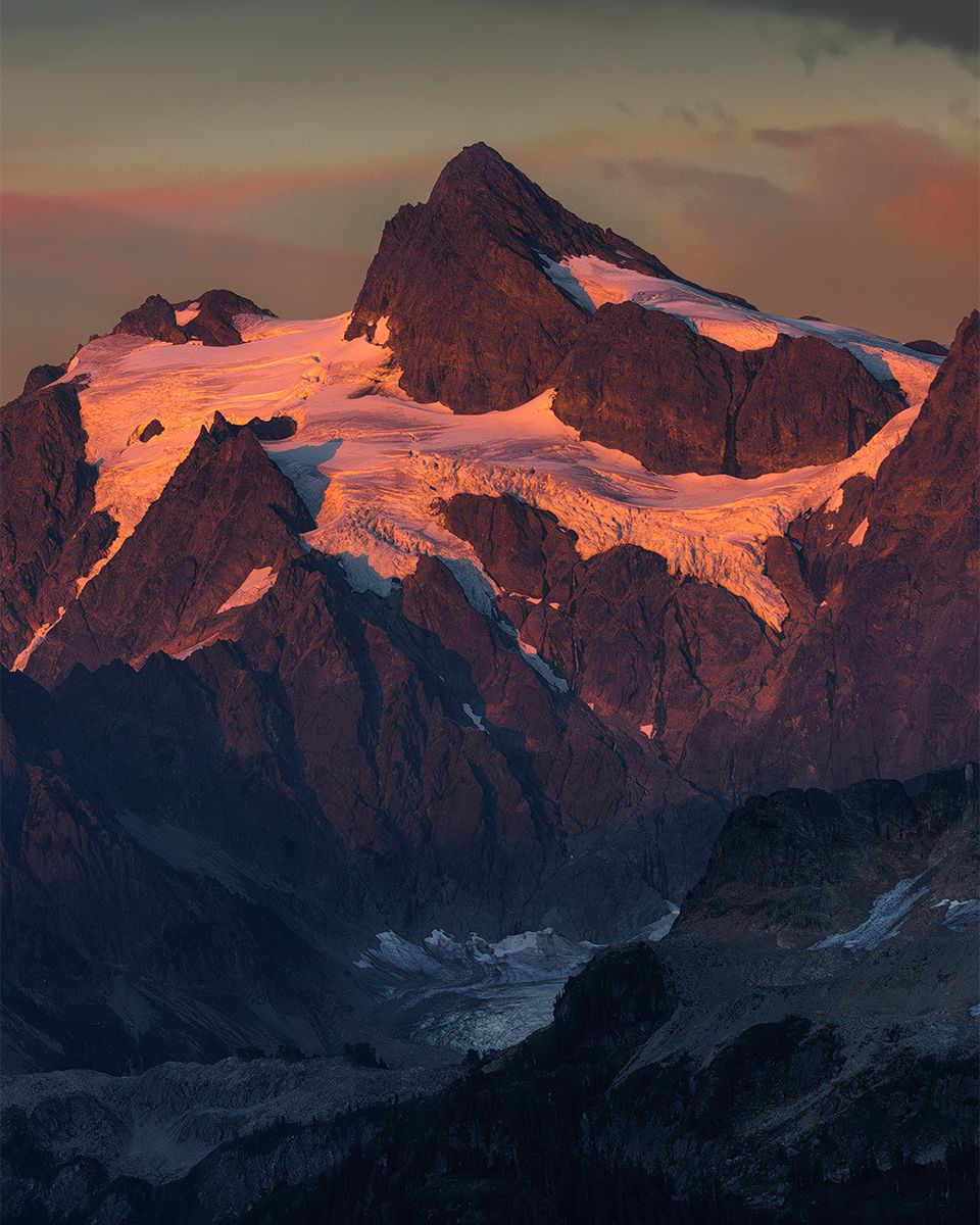 Dramatic sunset illuminates snow-capped mountain peaks and glaciers in a rugged alpine landscape.