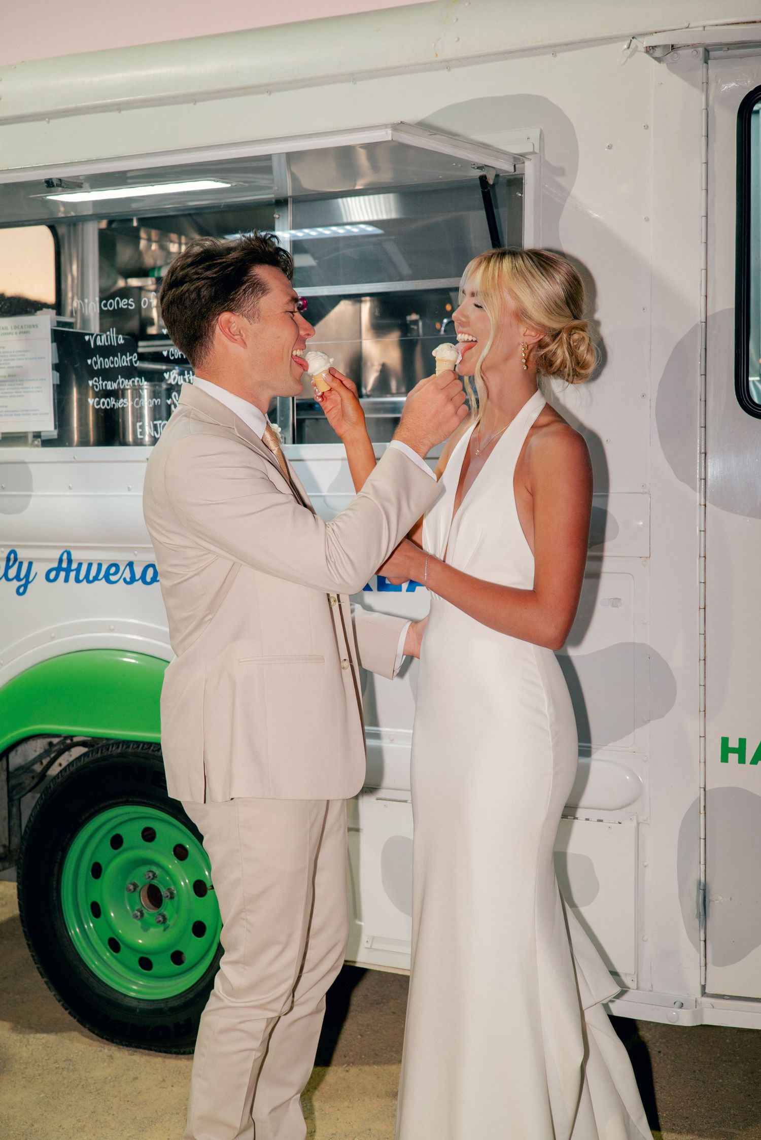 A couple in matching light colored formal attire playfully feed each other cake in front of a food truck.