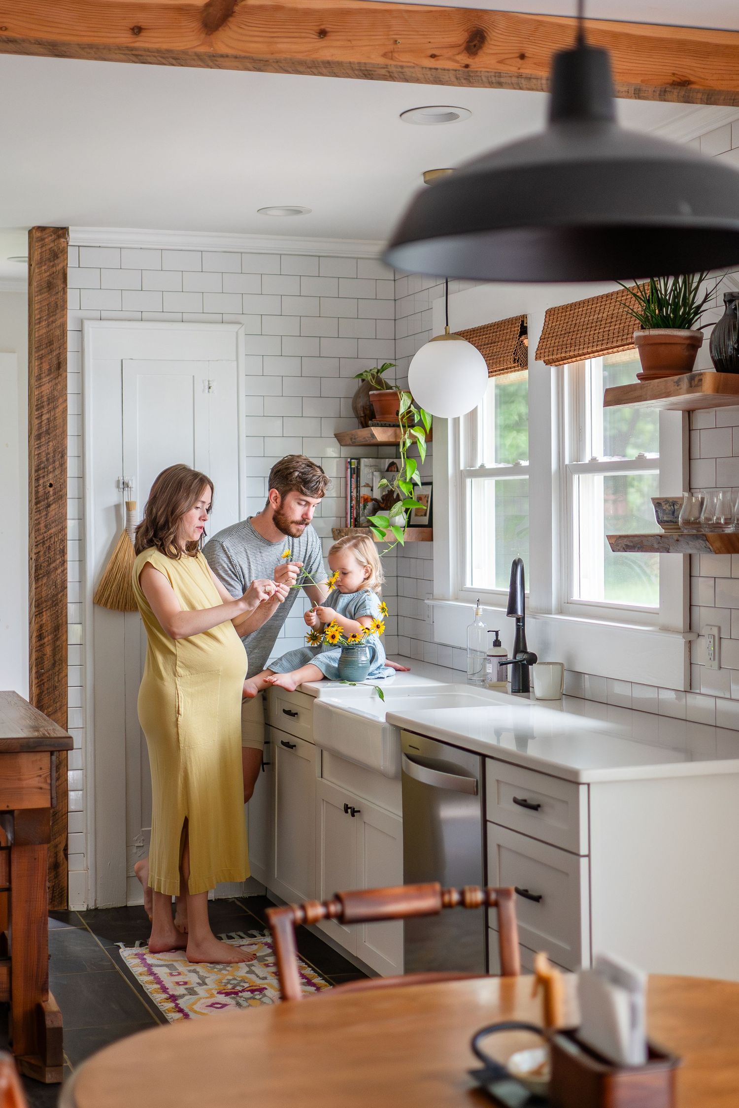 Modern farmhouse kitchen with white subway tile, wooden beams, and family gathered by sink under pendant lighting.