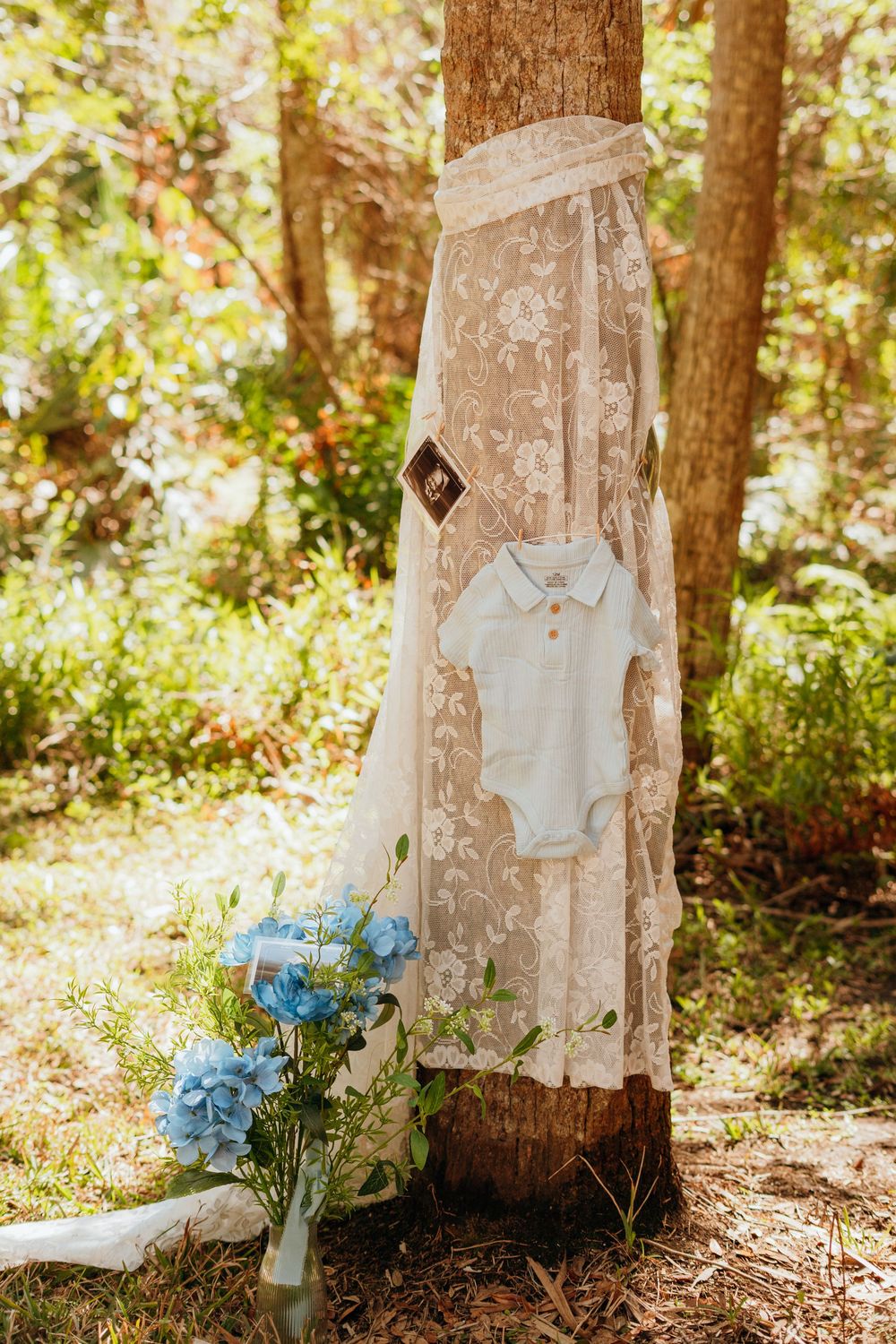 White lace wedding dress and blue flowers arranged decoratively on tree trunks in a sunlit forest setting.
