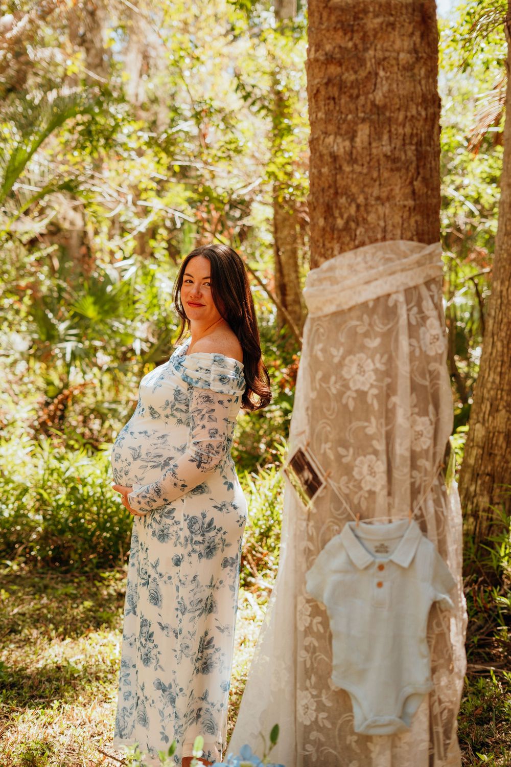 A woman in a floral blue and white dress poses in a sunlit forest setting with vintage clothing displayed on hangers.