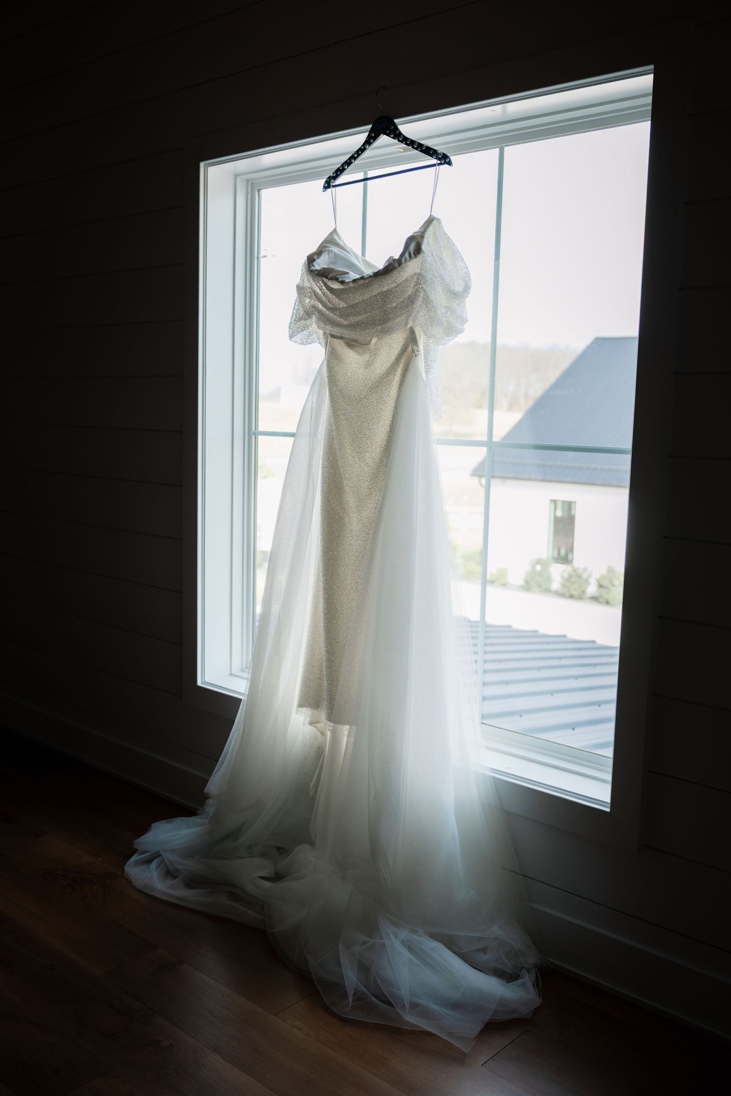 A white wedding dress hangs in silhouette against a bright window, creating an ethereal backlit glow.