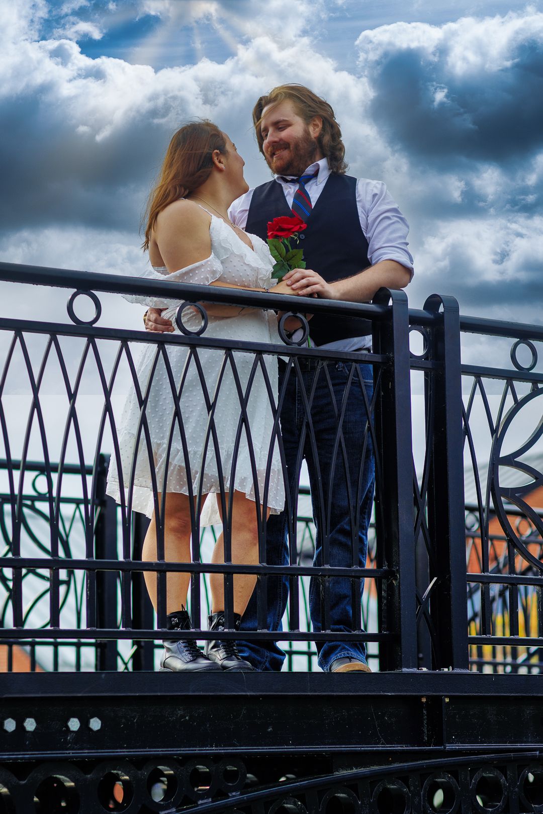 A couple shares a romantic moment on a balcony railing against a dramatic cloudy sky backdrop.