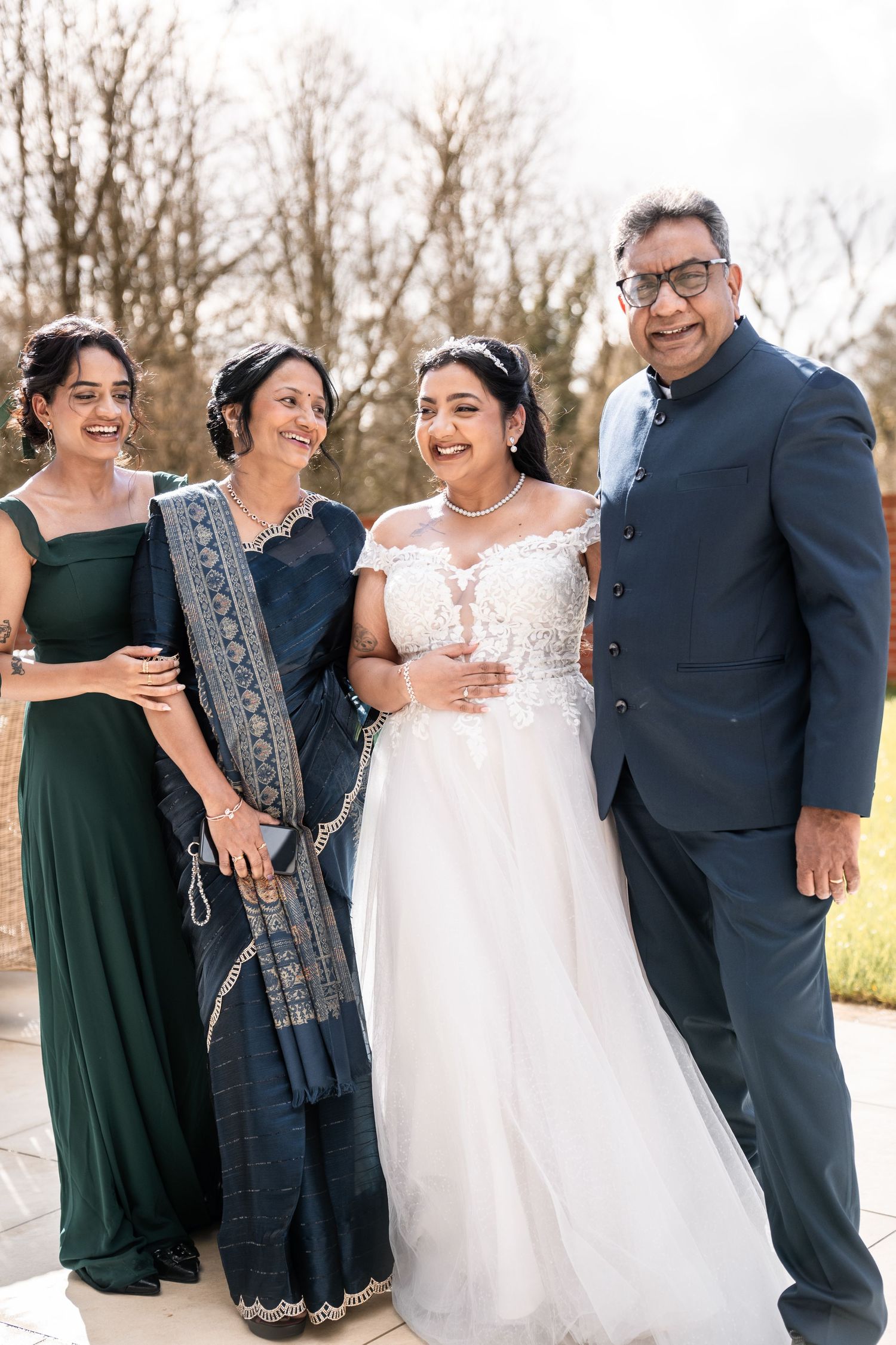 A wedding party laughs together outdoors wearing dark blue suits and green dresses with the bride in white.