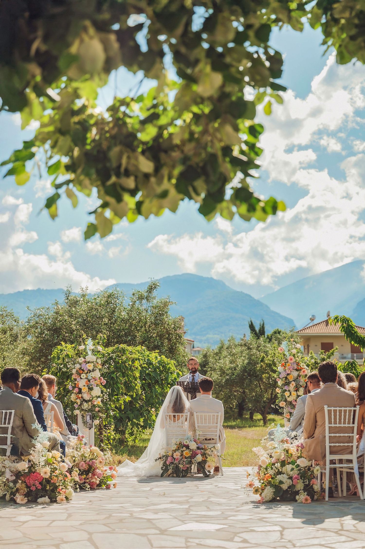 Outdoor wedding ceremony setup with white chairs and floral arrangements against a mountain backdrop on a sunny day.