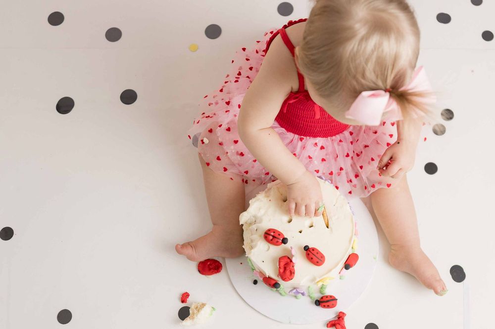 Baby in pink polka dot dress smashing and playing with a strawberry cake on polka dot background.
