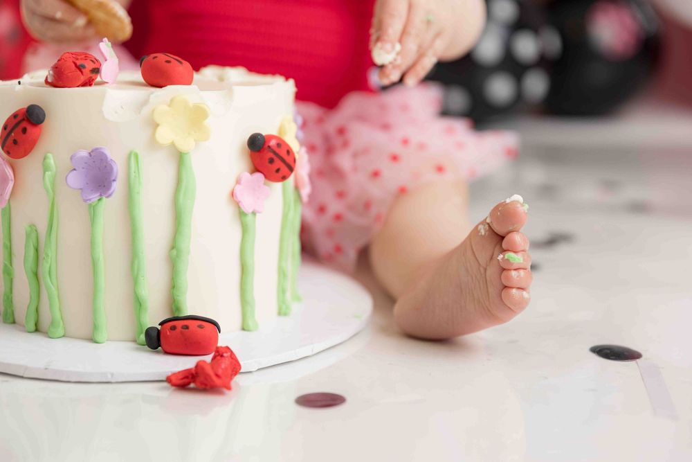 Small feet visible next to a white birthday cake decorated with ladybugs and green stems on a light surface.