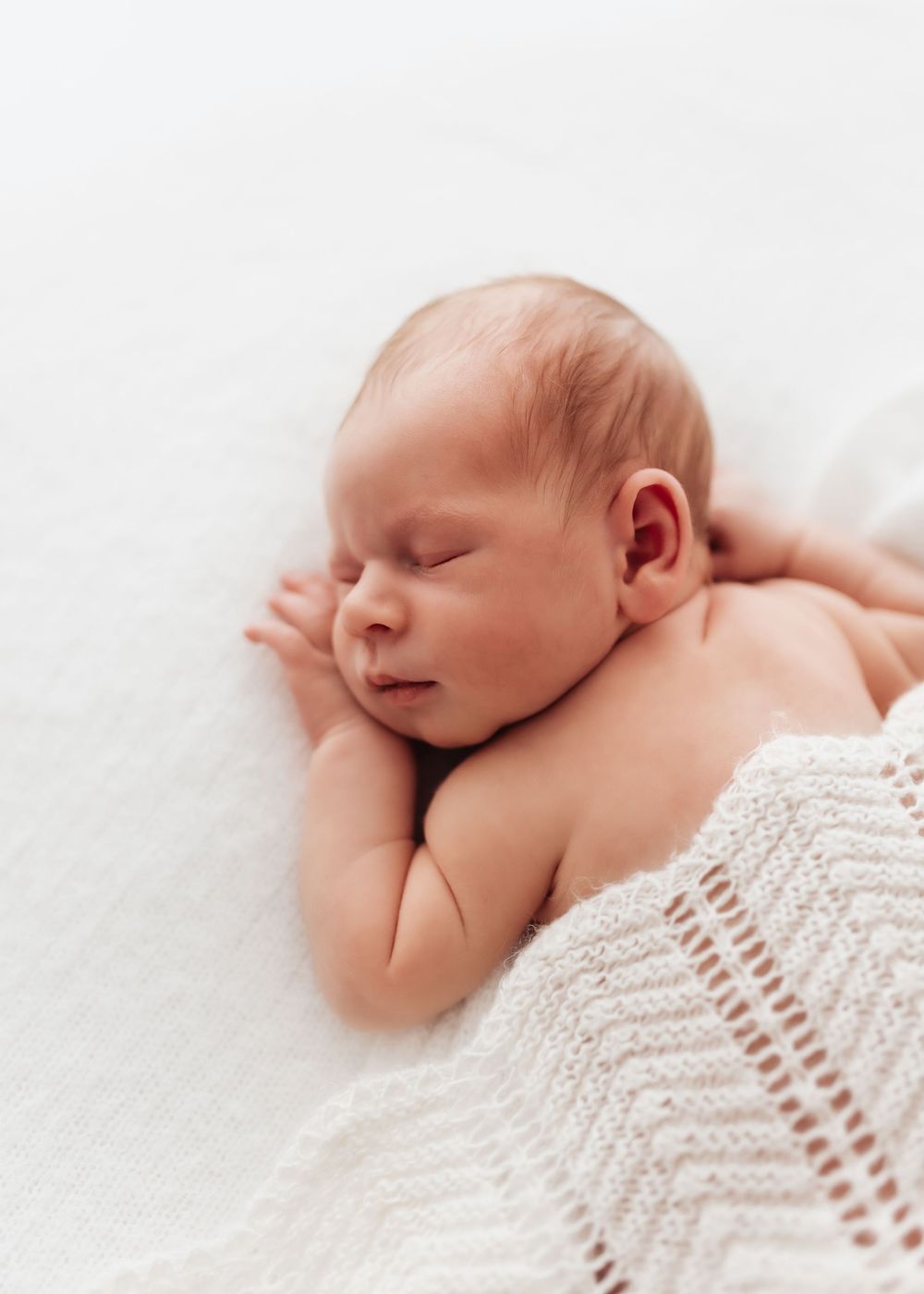 A peaceful sleeping newborn wrapped in a delicate white knit blanket lies on a soft white surface.