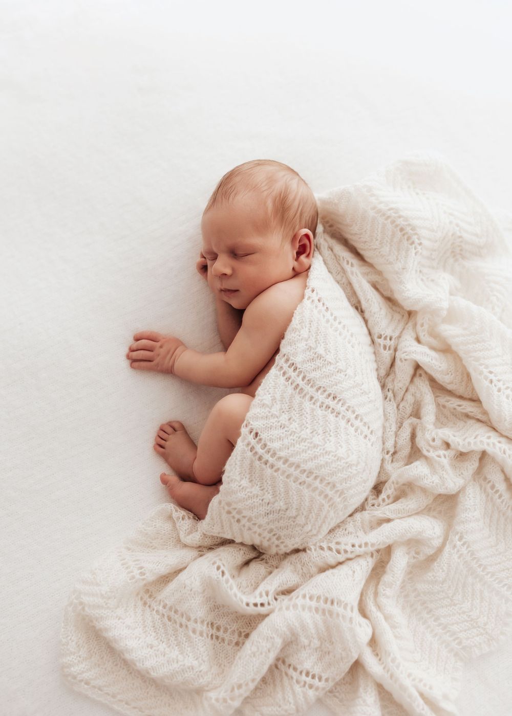 Sleeping newborn baby wrapped in a cozy white knitted blanket against light background.