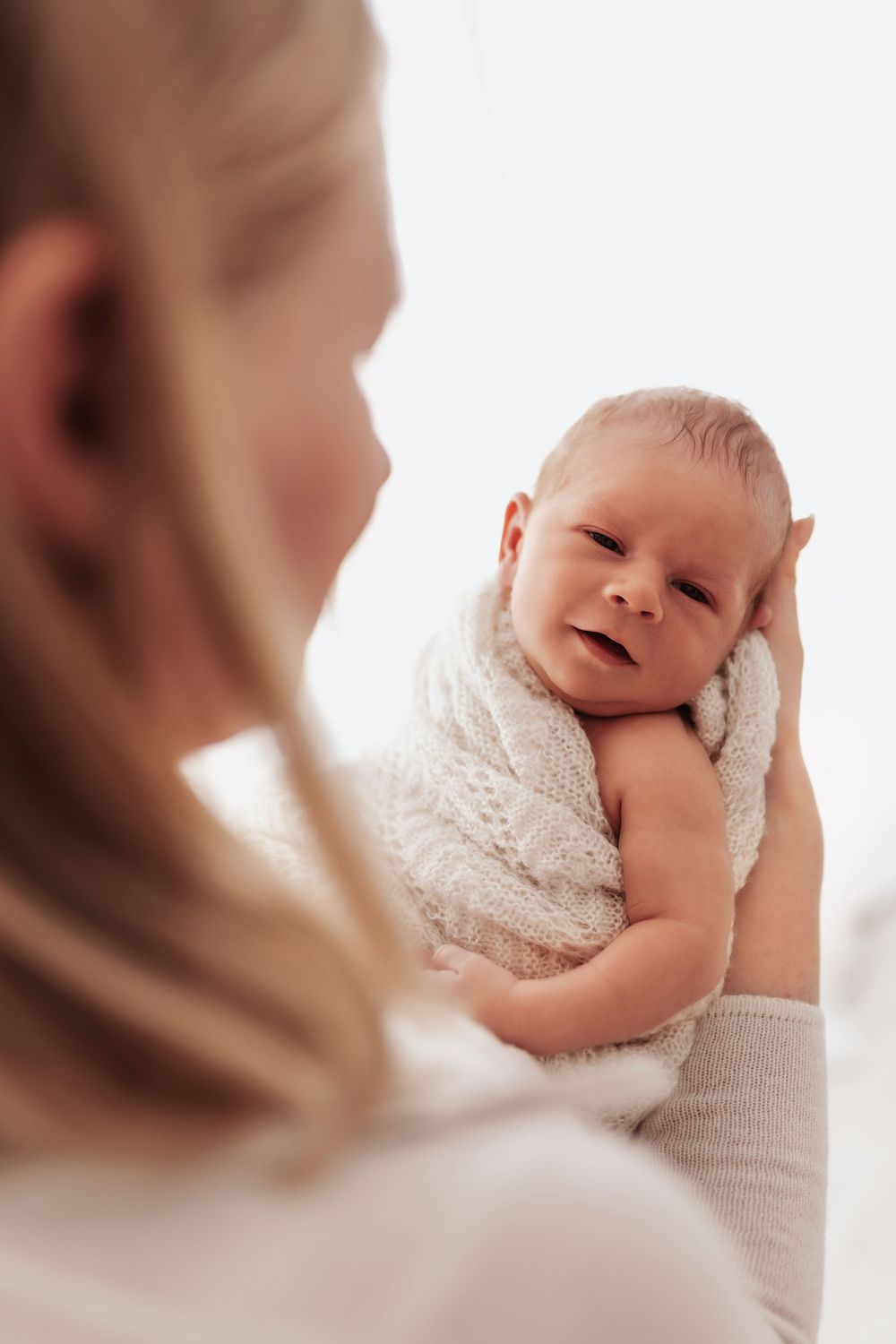 Baby wrapped in cream colored blanket held gently against light background.