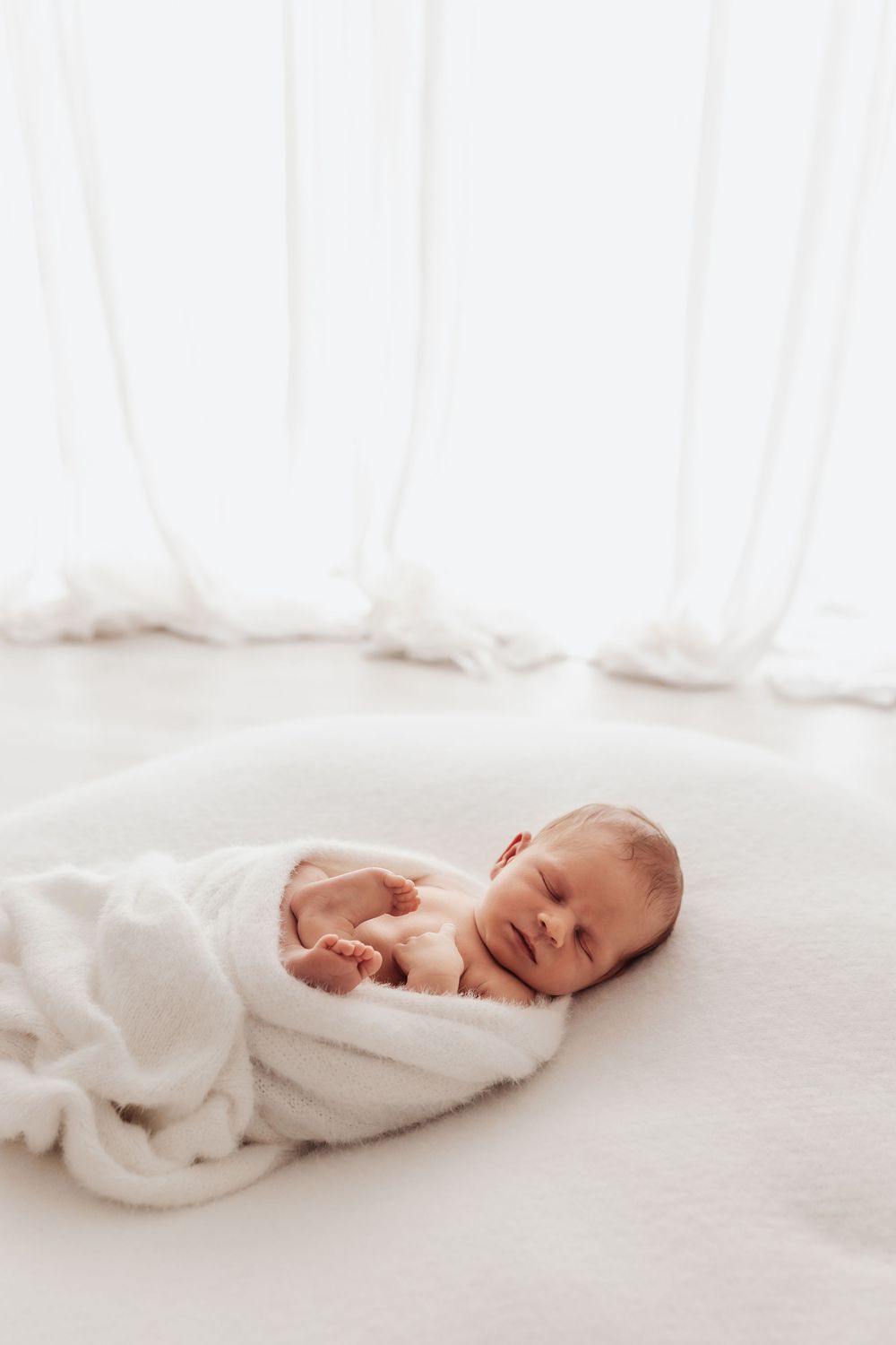 Peaceful newborn baby wrapped in soft white blanket resting on white bedding in bright minimalist setting.