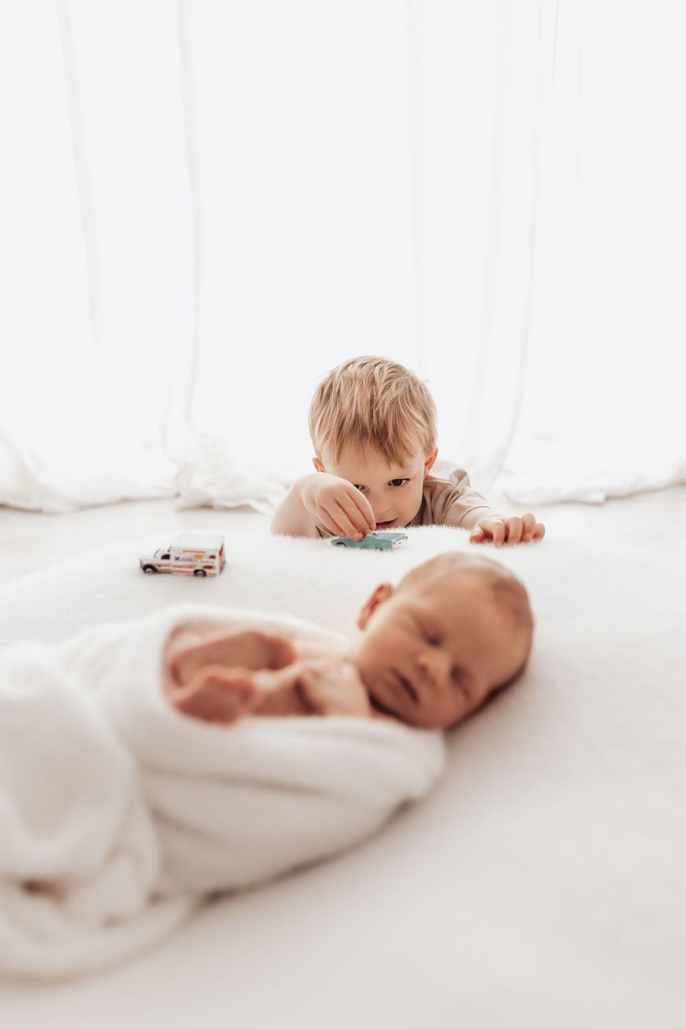 A toddler peeks at a sleeping newborn baby wrapped in white swaddling on a light colored surface.