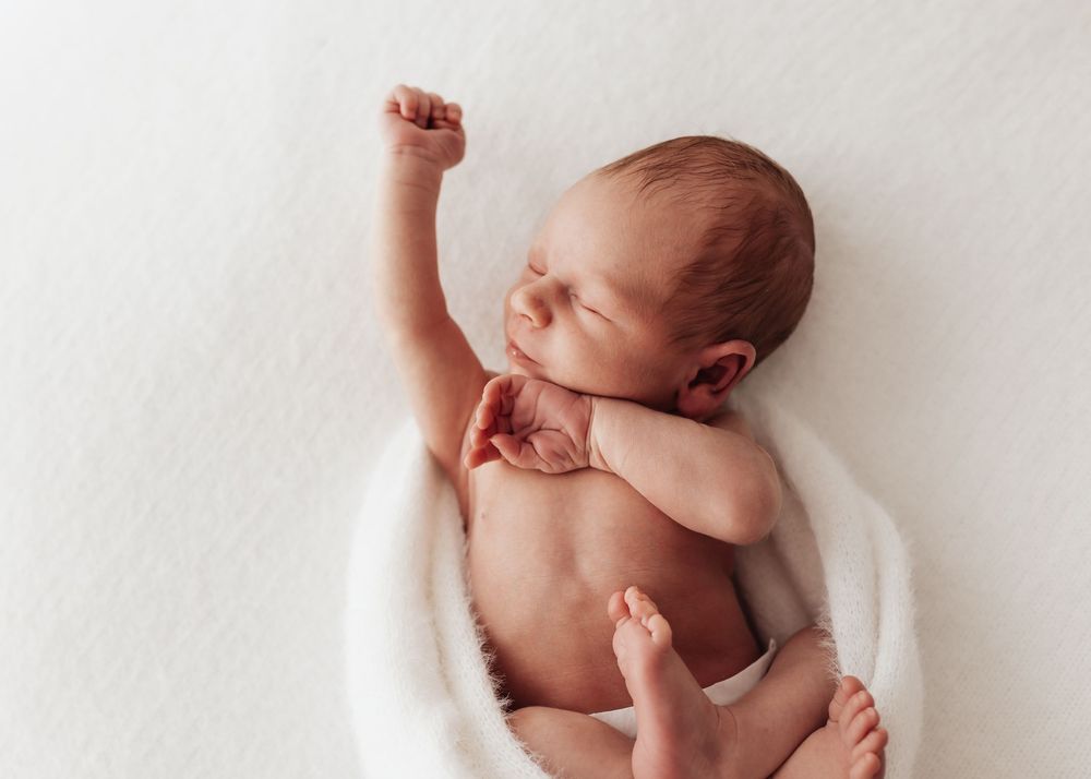 A newborn baby lying on a white blanket stretches their tiny arms and legs during a professional photo session.