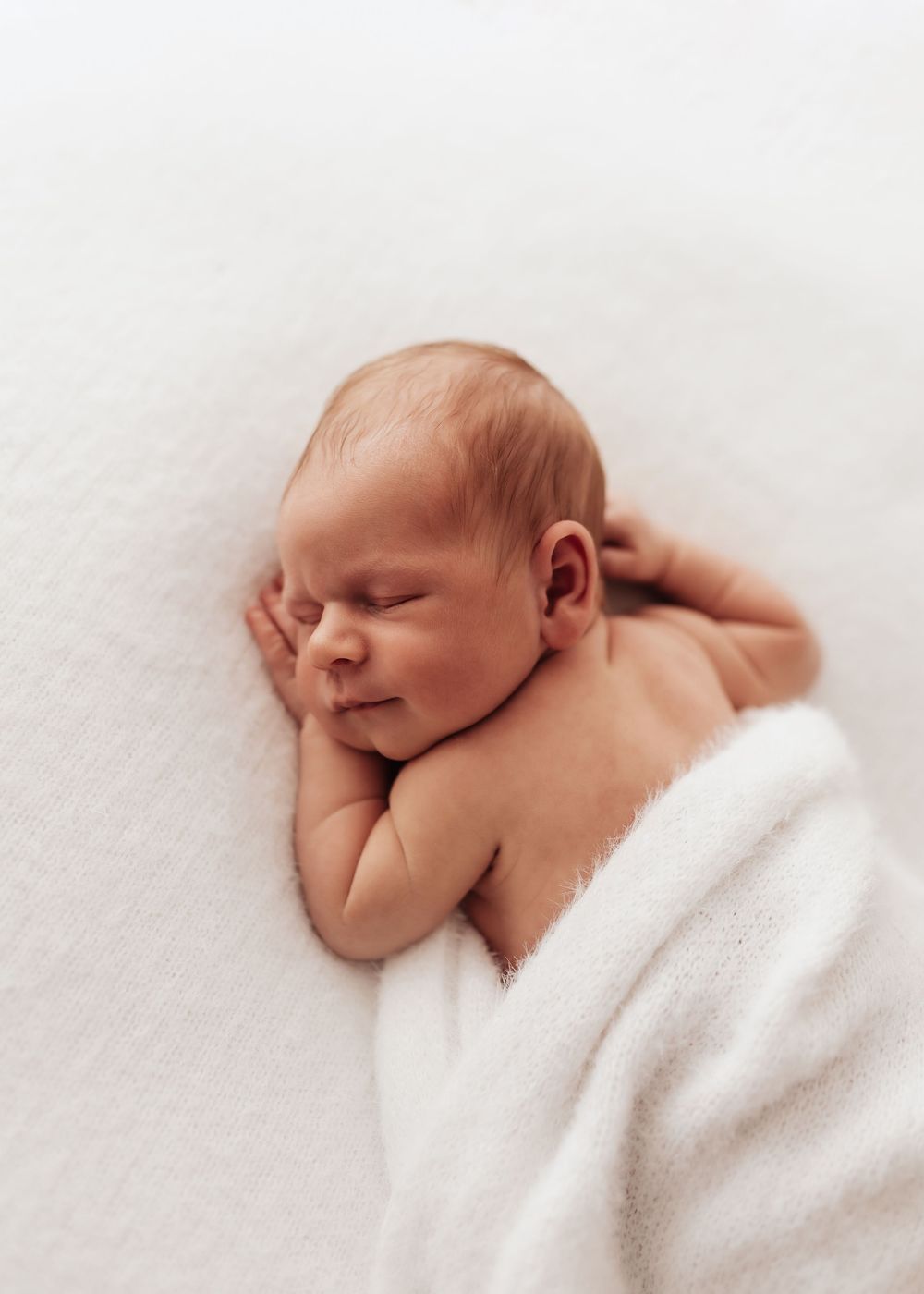 Peaceful newborn baby wrapped in white blanket sleeping on light background.