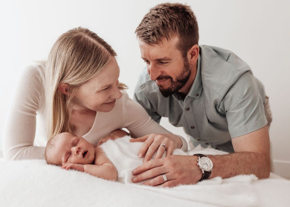 Parents lovingly gazing at their newborn baby while lying together on white bedding.