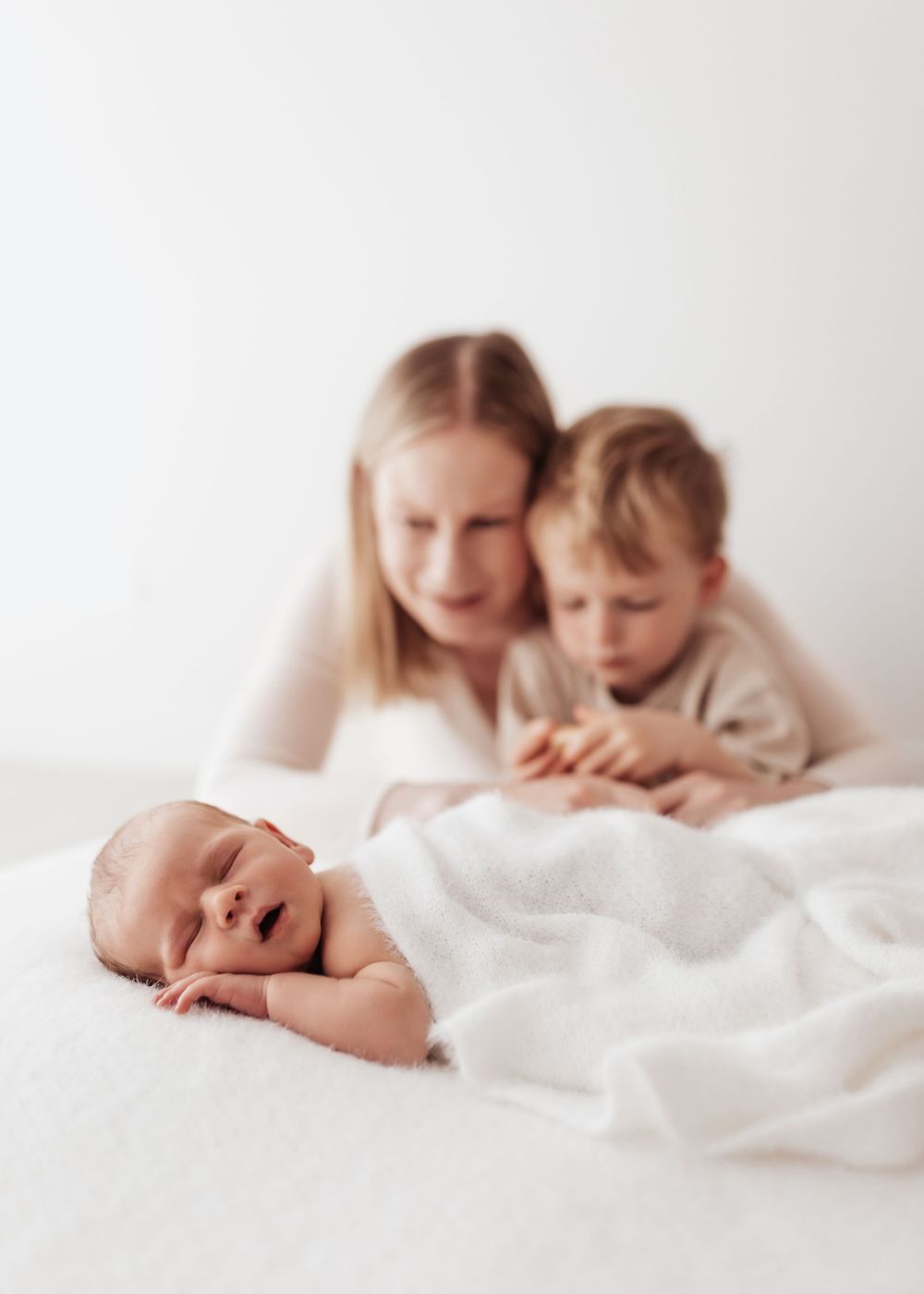 Tender moment captured in soft light as siblings gaze at sleeping newborn on white bedding.
