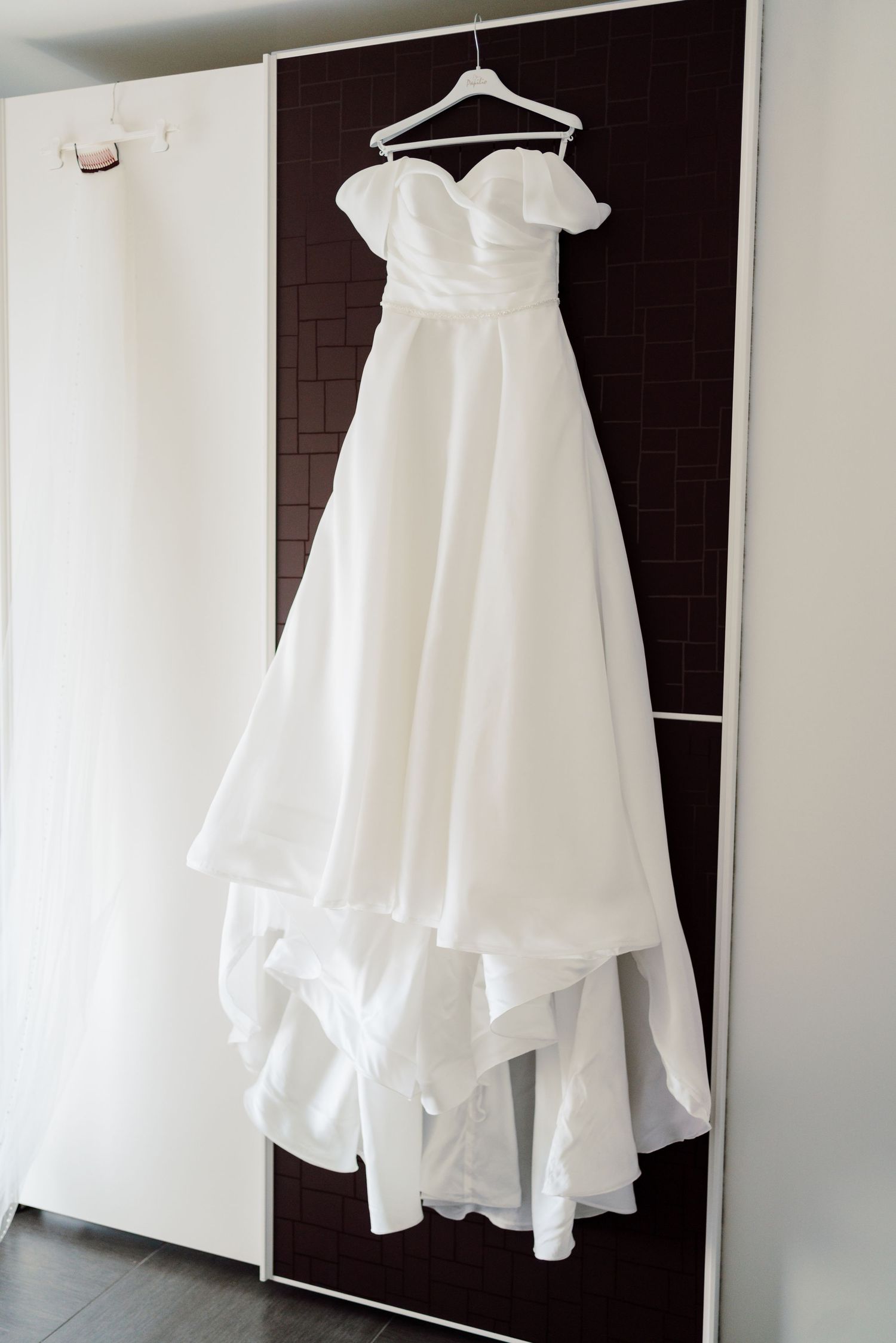 White off-shoulder A-line wedding dress hanging on a hanger against a dark wardrobe door.