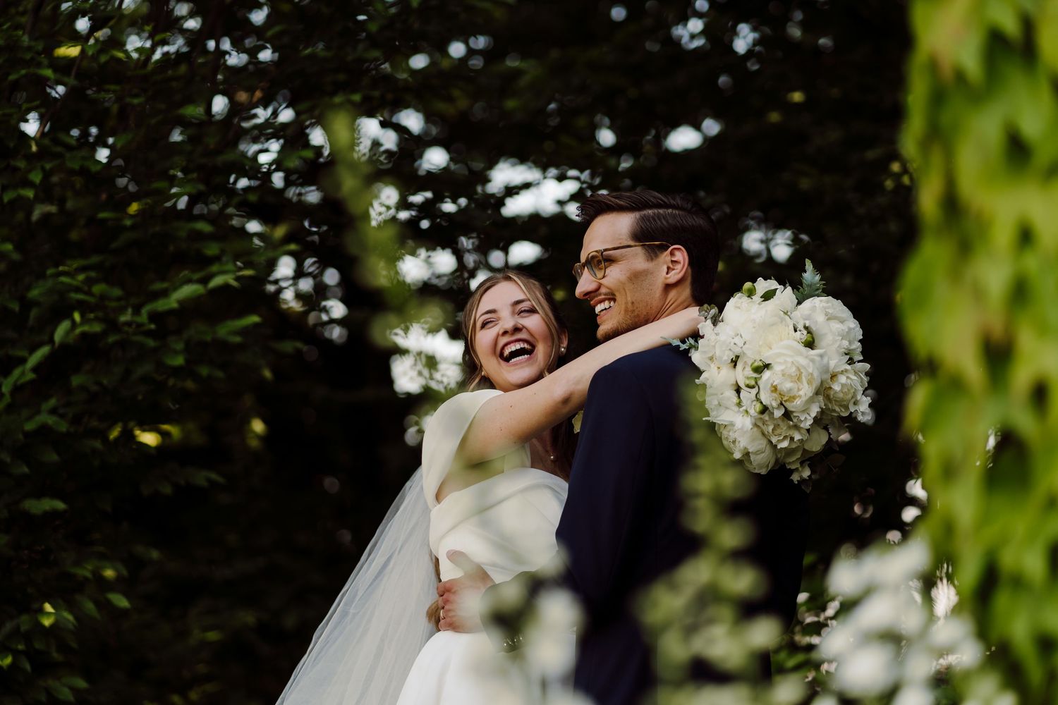 Joyful bride and groom laughing together outdoors with lush green trees behind them.