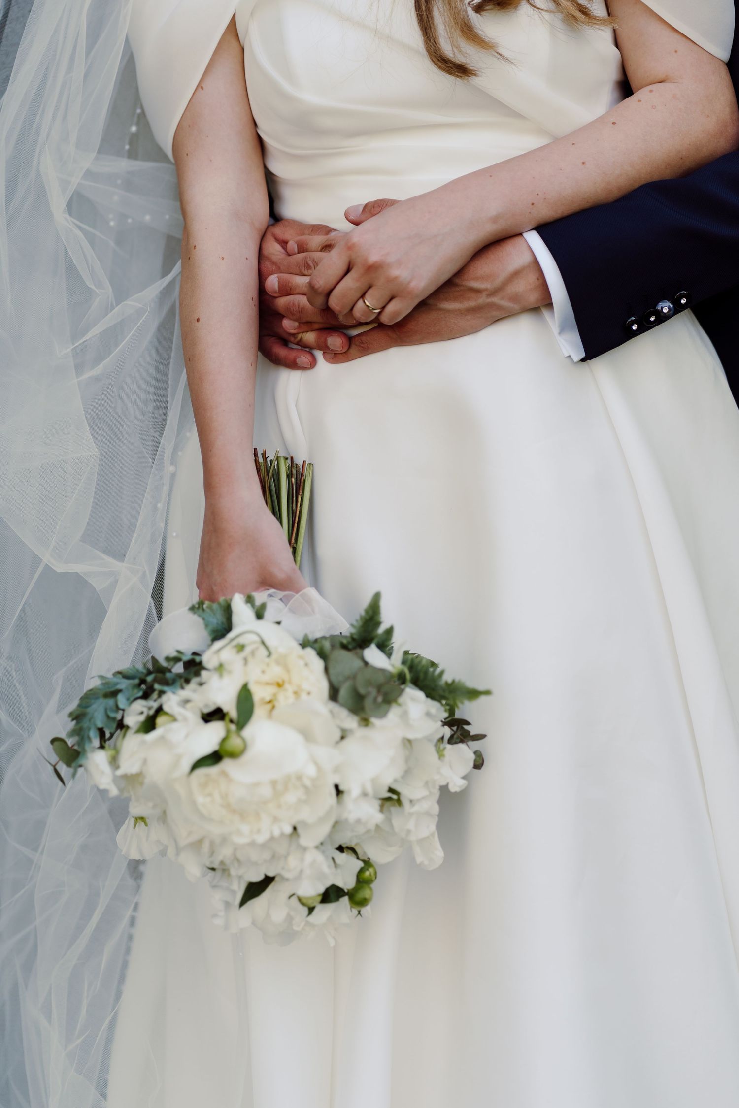 Bride and groom holding hands at waist, bride holding white peony bouquet in elegant ivory wedding gown.
