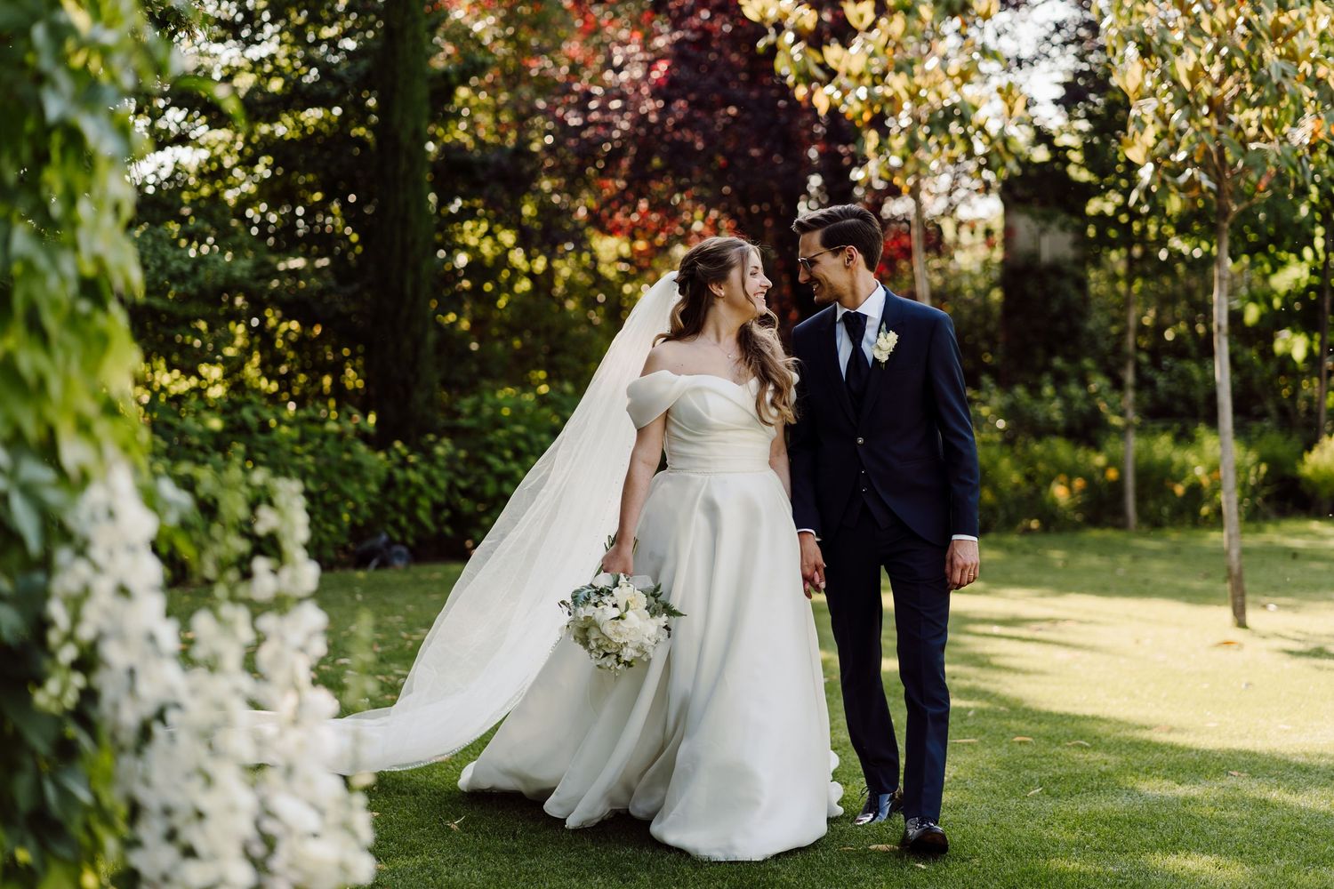 Bride and groom walking in lush garden, bride in off-shoulder gown with cathedral veil holding white bouquet.