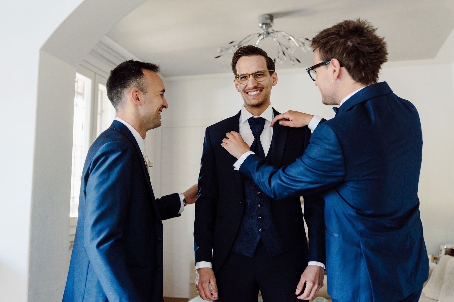 Three groomsmen in navy suits helping each other prepare for a wedding ceremony indoors.