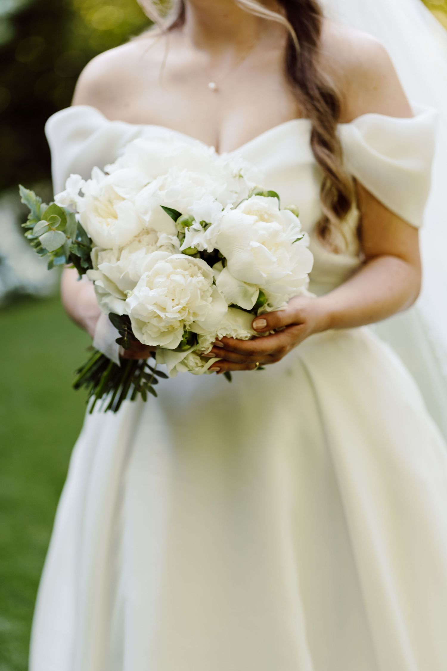 Bride in off-shoulder ivory gown holding lush white peony and greenery wedding bouquet outdoors.