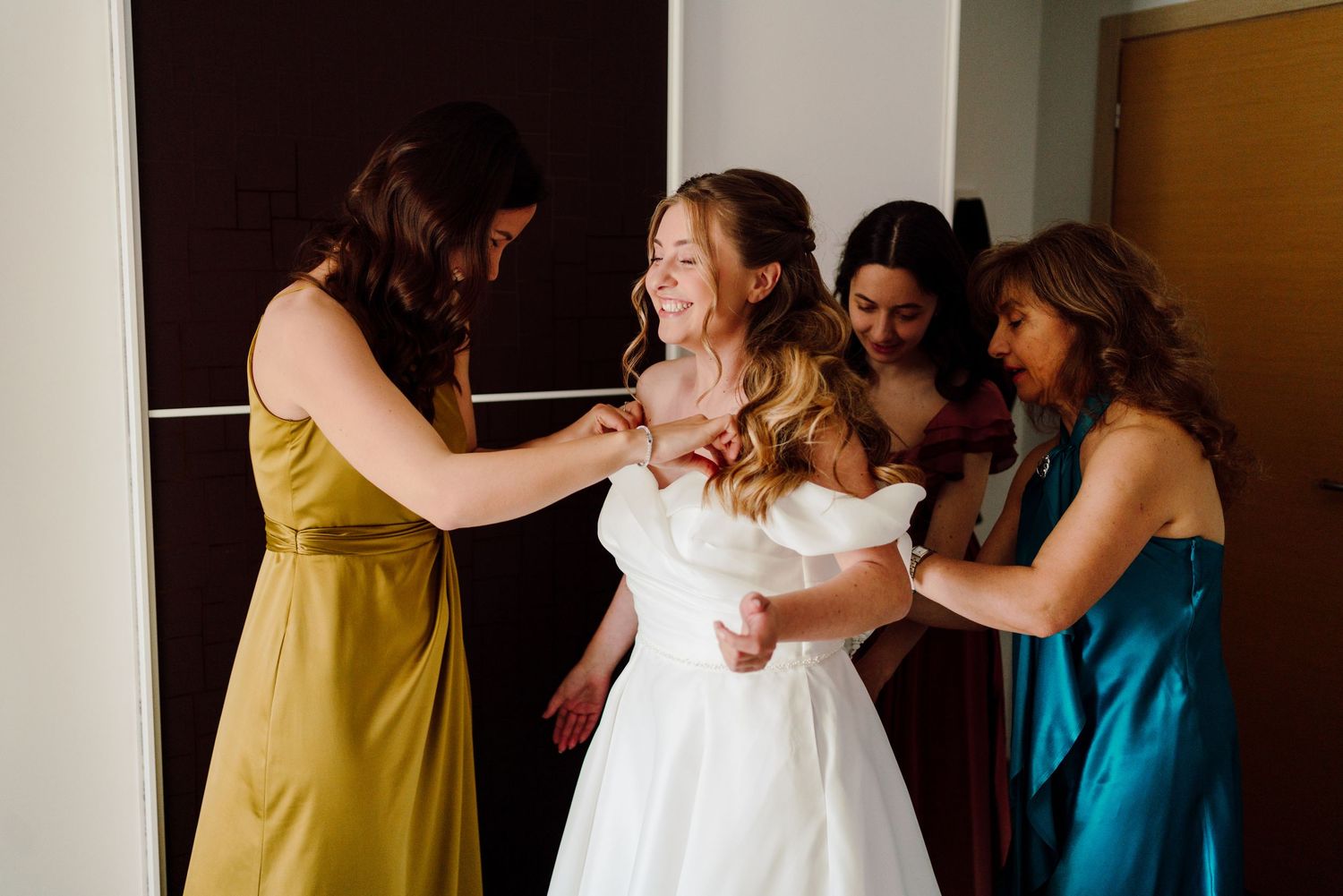 Bride smiling as three bridesmaids in colorful dresses help her into her white wedding gown.
