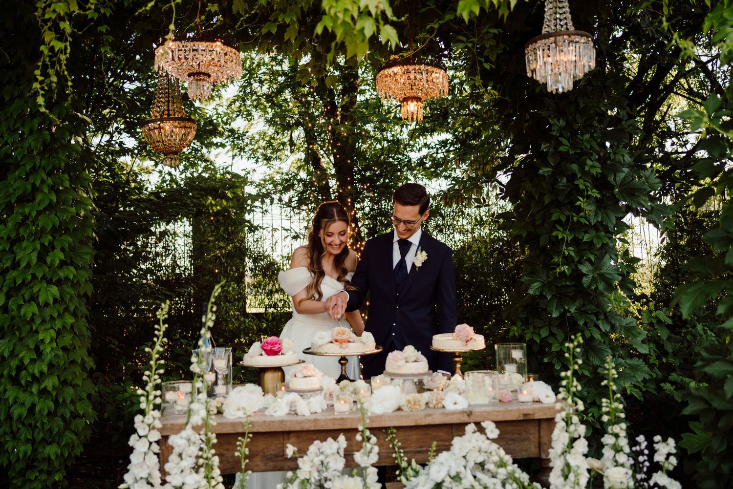 Bride and groom cutting wedding cake at floral dessert table under crystal chandeliers in garden setting.