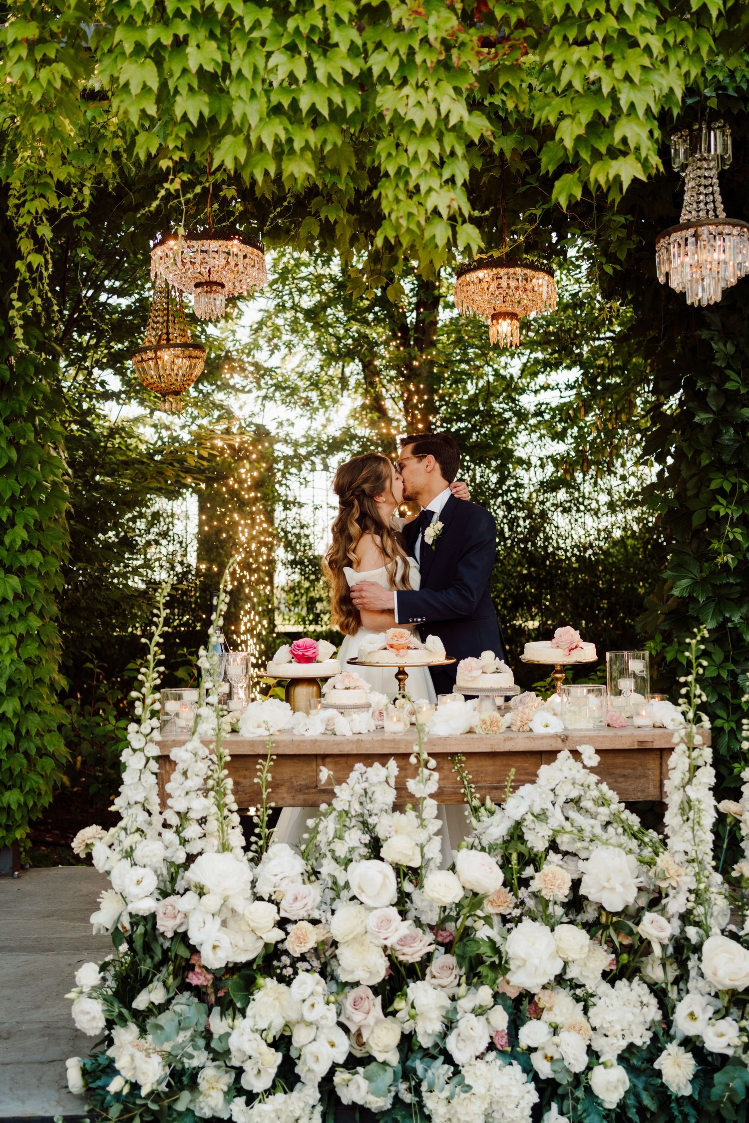 Bride and groom kissing behind elegant dessert table adorned with white flowers under crystal chandeliers.