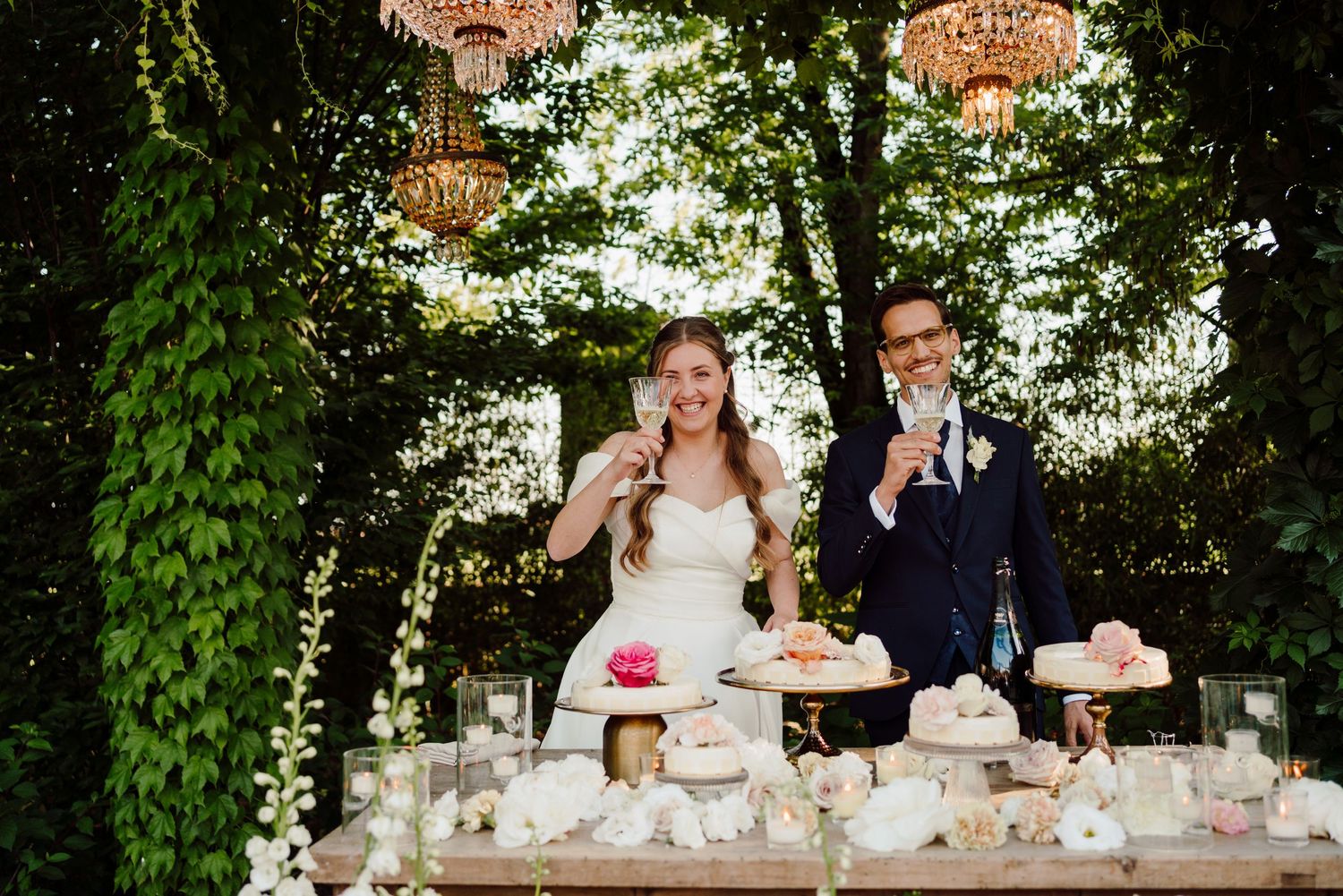 Happy bride and groom toasting beside an elegant dessert table with chandeliers and greenery backdrop.