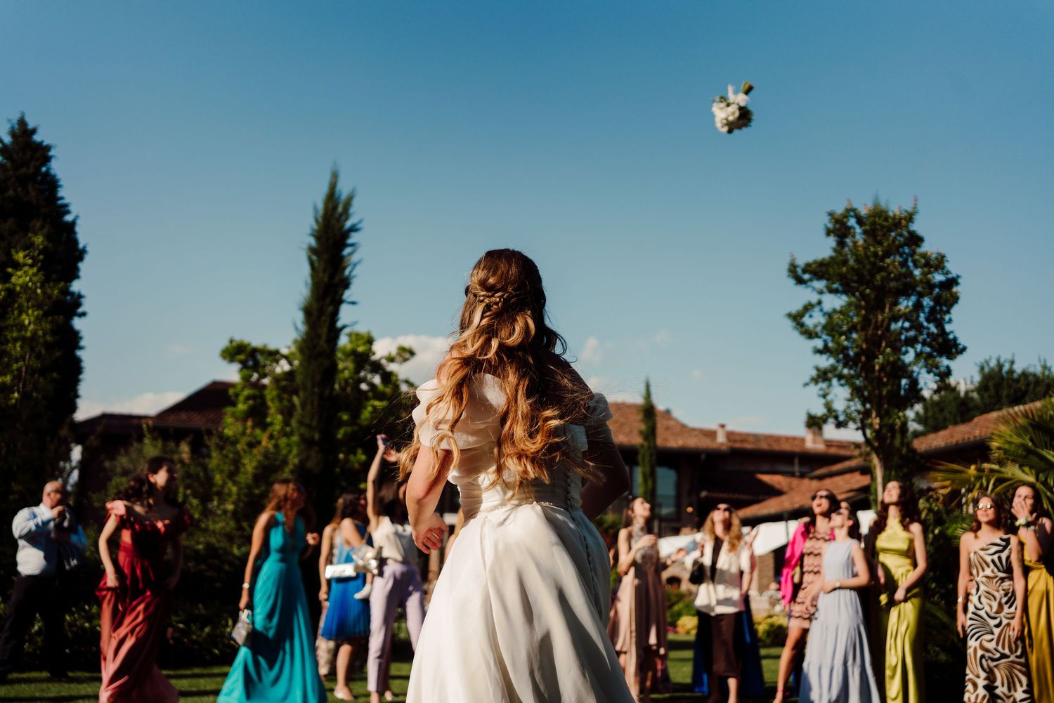 Bride tossing wedding bouquet to guests gathered on sunny outdoor lawn during wedding reception.