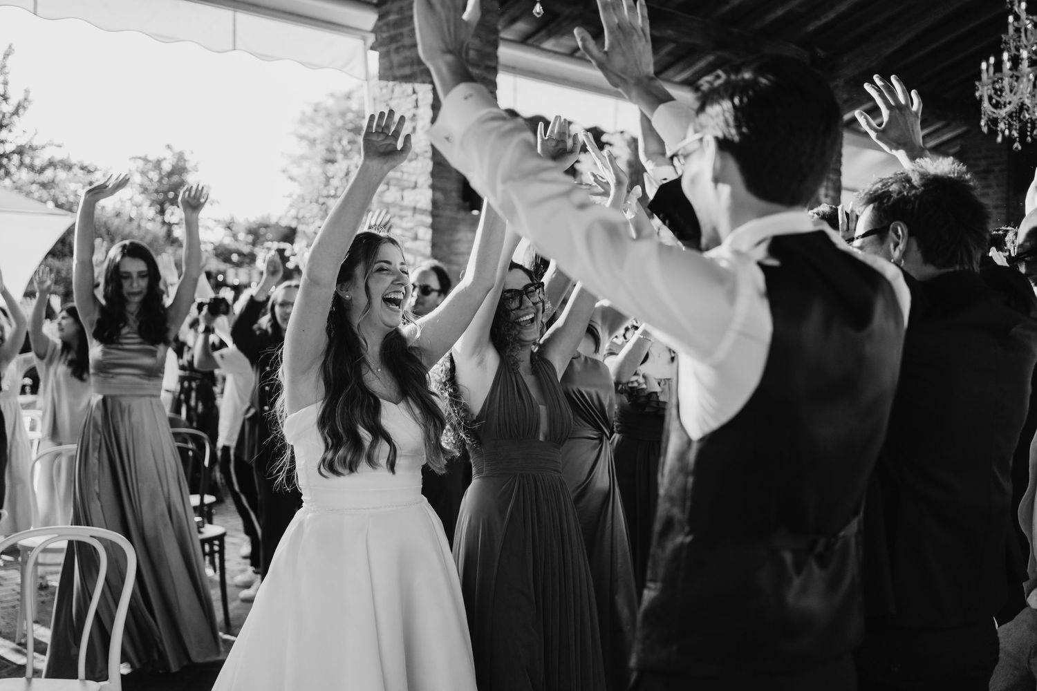 Black and white photo of bride in white gown celebrating with cheering wedding guests at outdoor reception.
