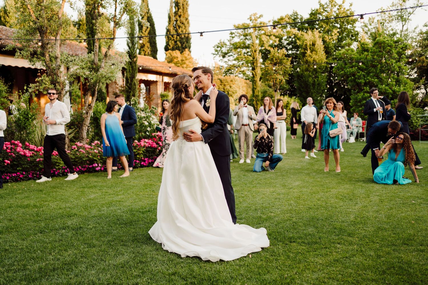 Bride and groom sharing first dance kiss on lush garden lawn surrounded by wedding guests.