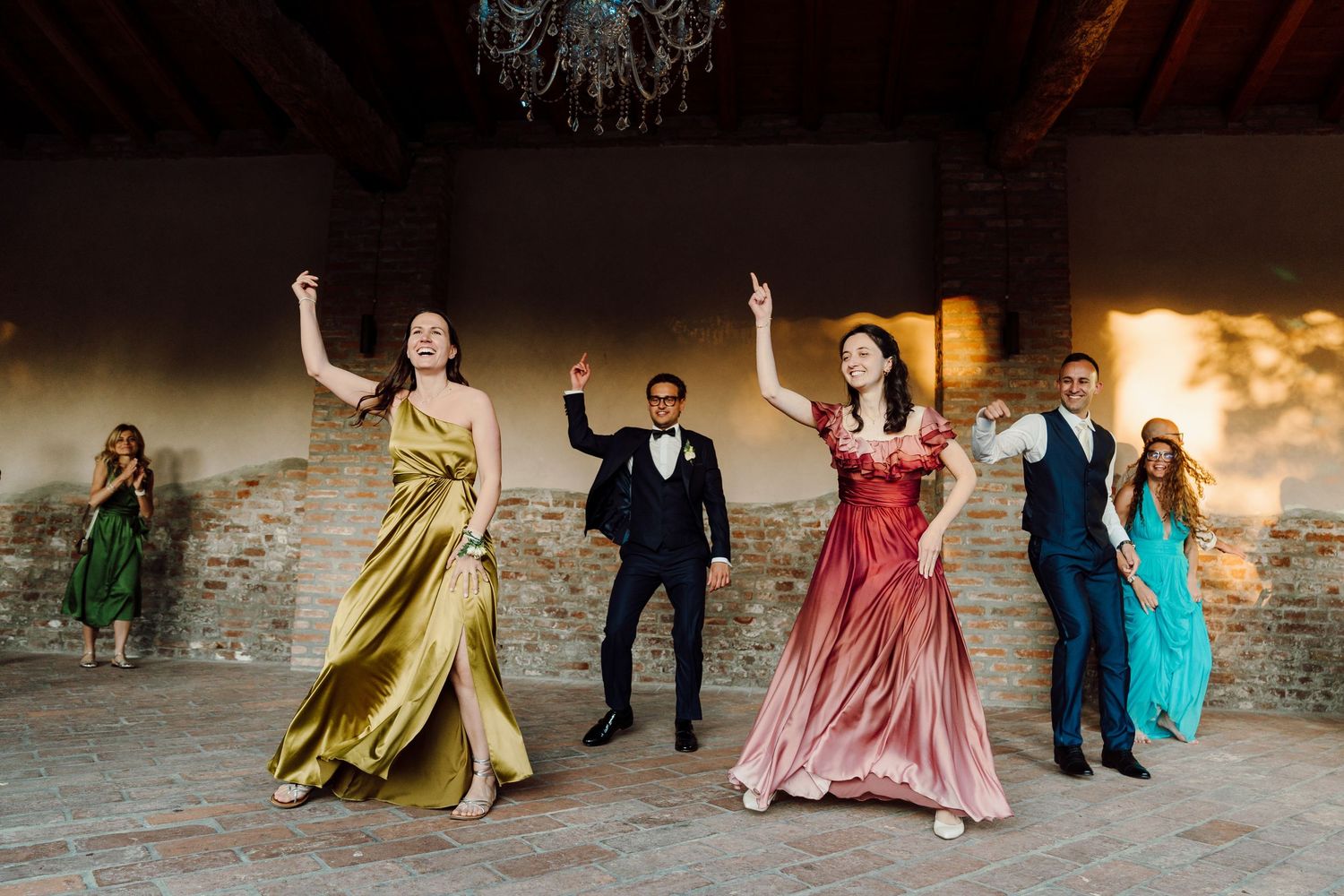Colorful wedding guests in elegant gowns dancing joyfully at an outdoor reception venue with stone walls.