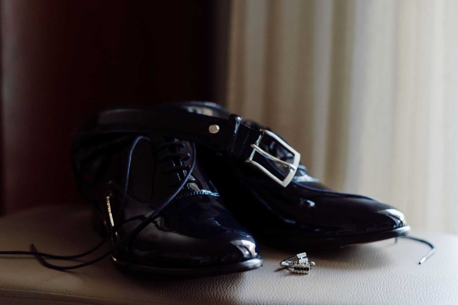 Shiny black patent leather groom's dress shoes with a belt and cufflinks on a surface.