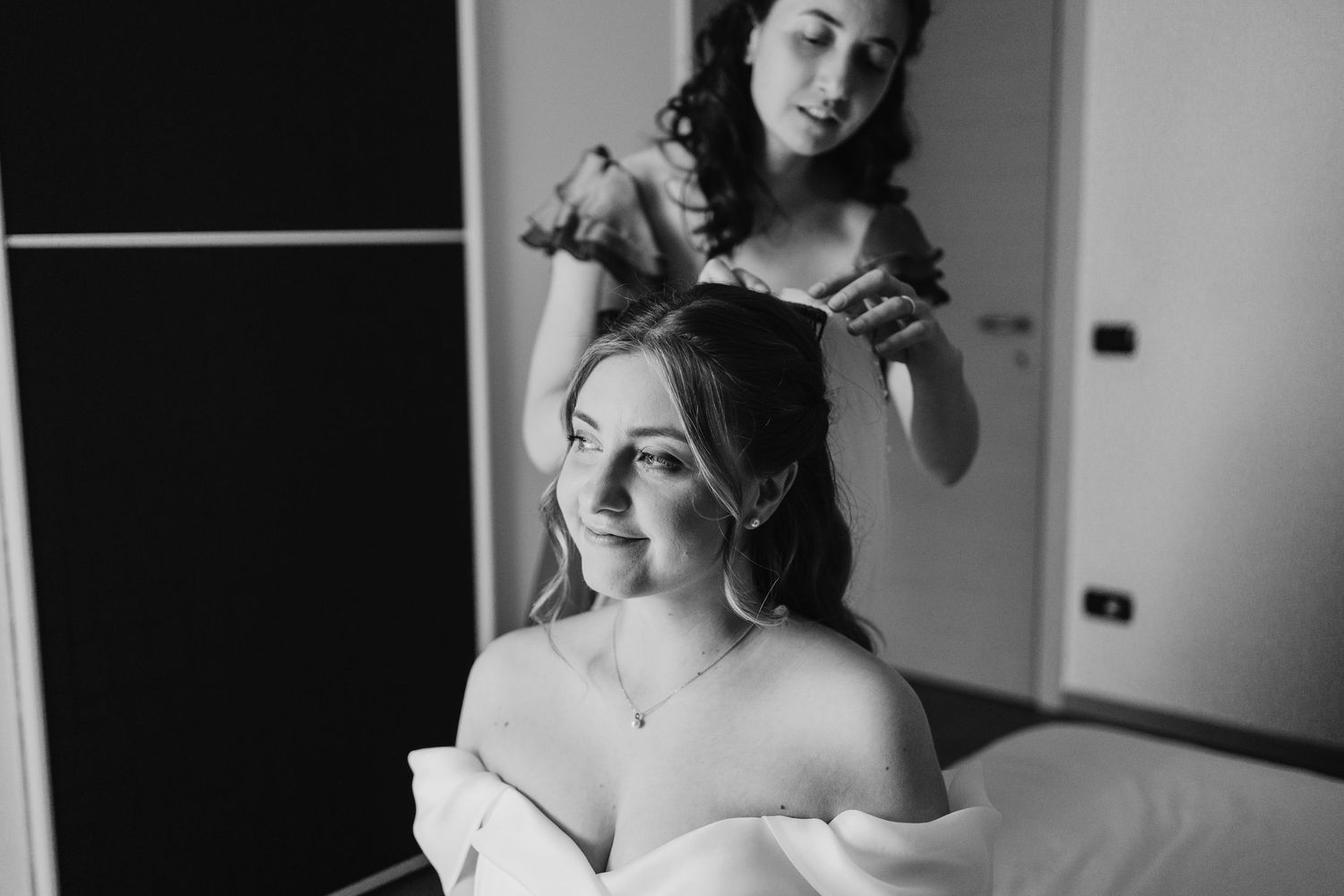Black and white photo of a hairstylist styling a smiling bride's hair before the wedding.